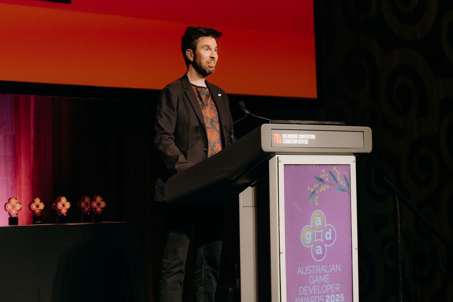 A person presenting on stage at a tech conference with digital displays showing tech-related graphics in the background.