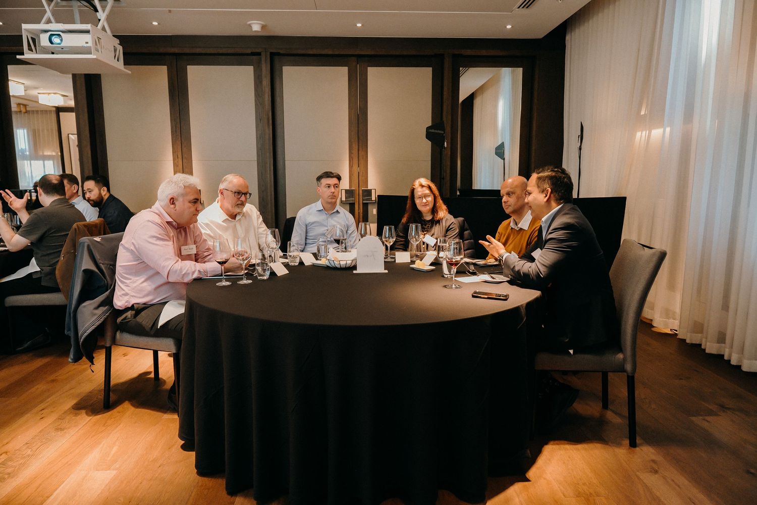 Business meeting attendees gather around tables in a modern conference room with warm lighting.