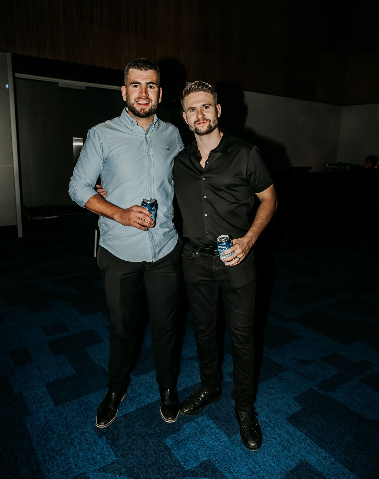 Two men in business casual attire pose together on a blue carpet in a dimly lit setting.