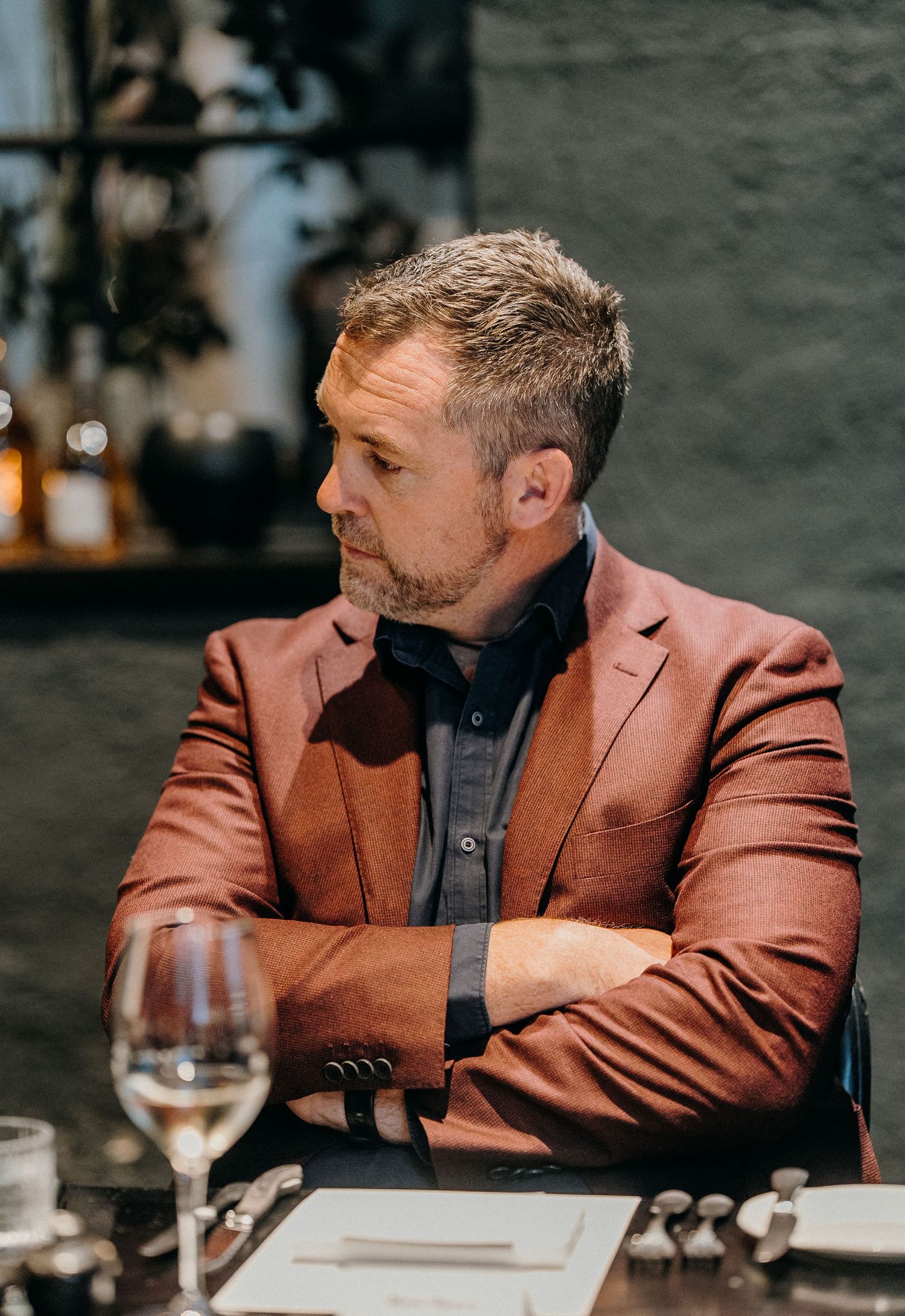 Man in brown blazer and dark shirt sits at restaurant table with wine glass and menu.