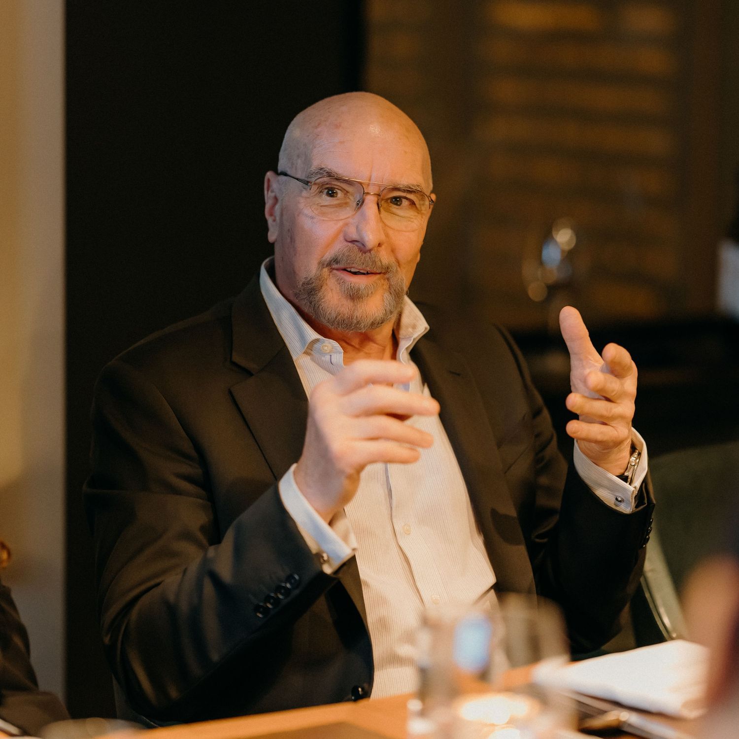 A person in a suit sits at a table in a dimly lit room, gesturing thoughtfully during a conversation.