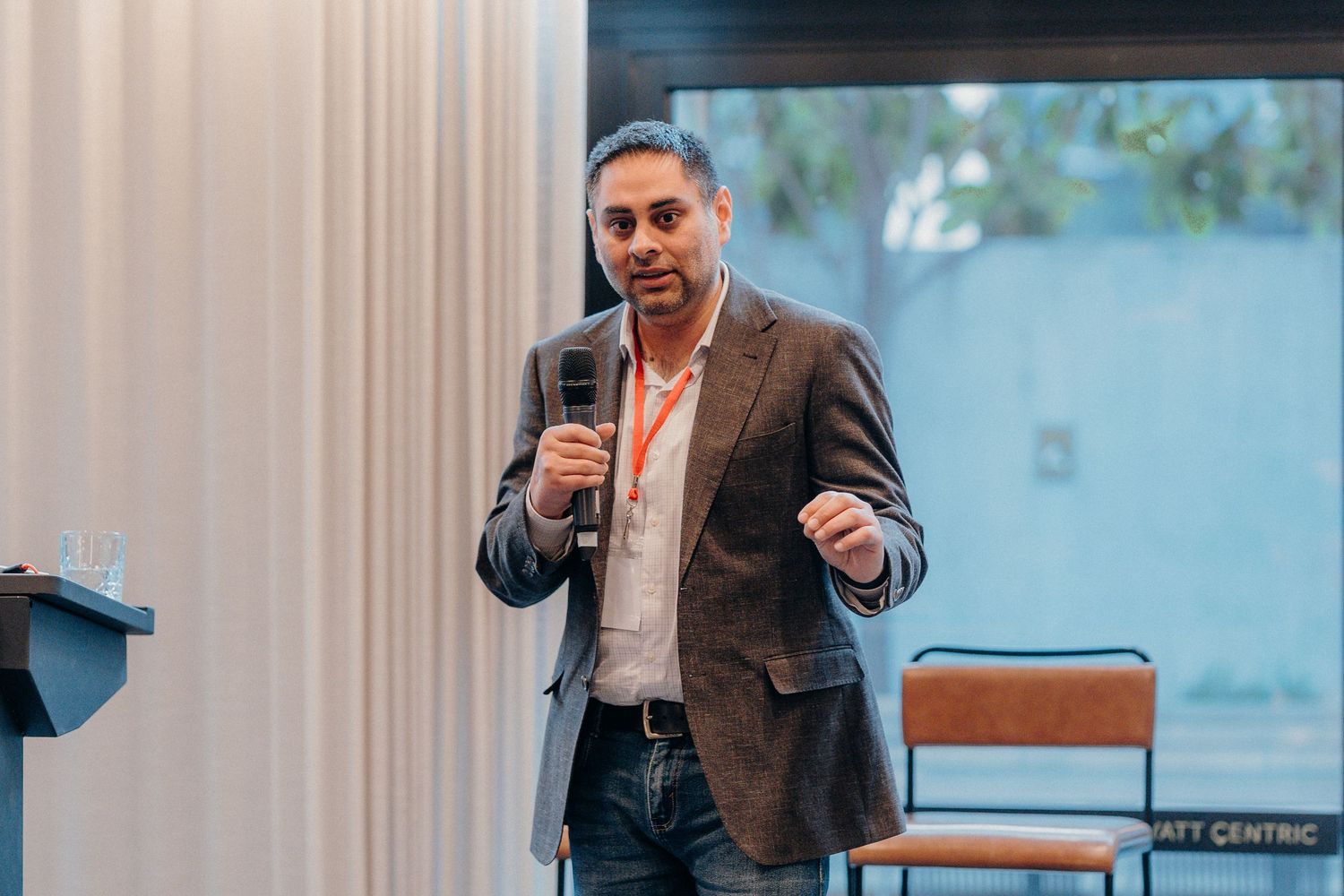 A speaker wearing a brown jacket presents to an audience during a business conference or seminar.