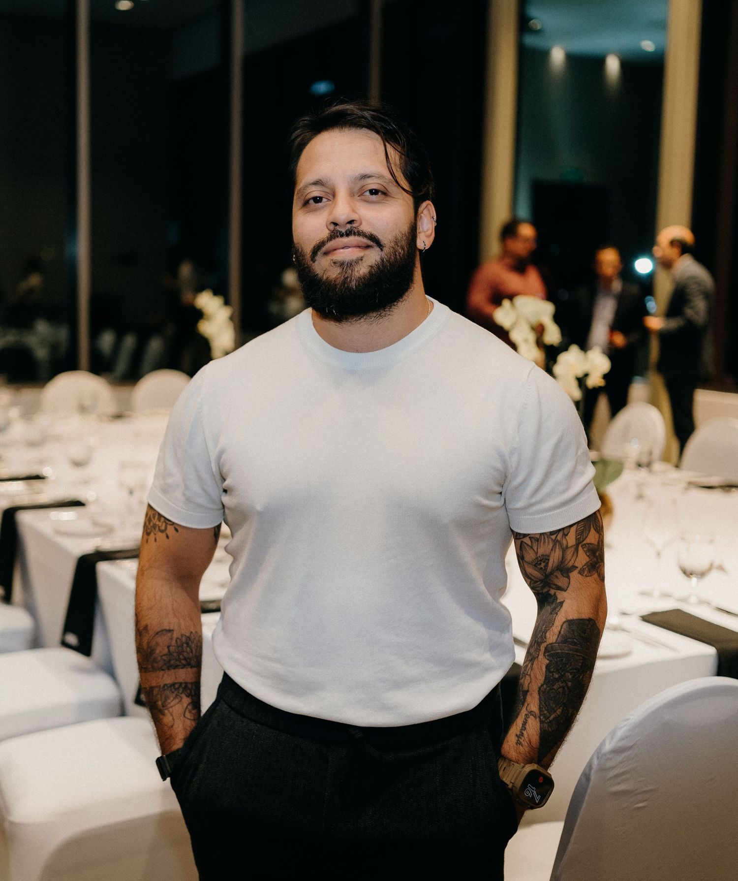 A person in a white t-shirt with tattoos sits at a formal dining event with white tablecloths visible in background.