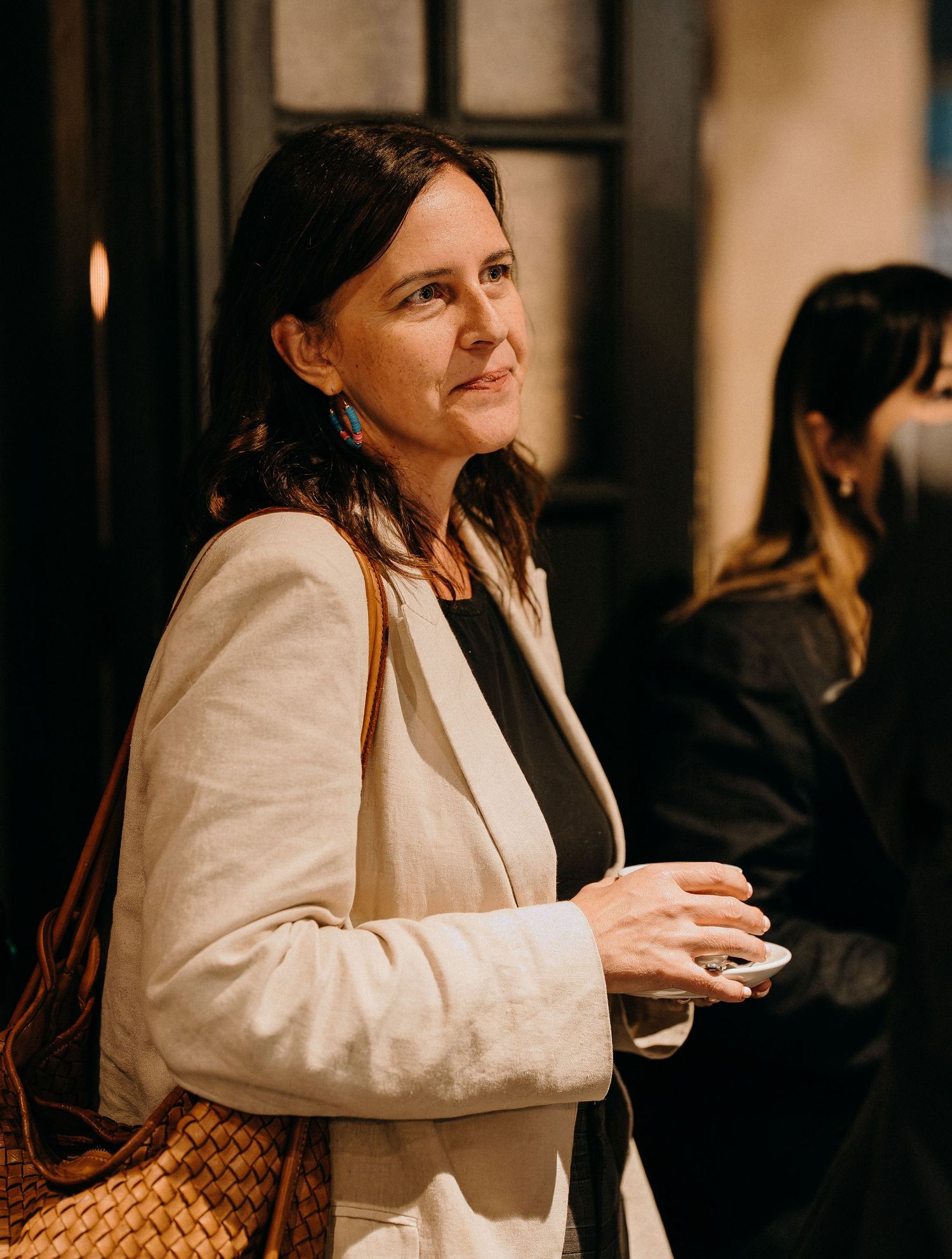 Woman in cream blazer and brown woven bag looks to the side while standing in dimly lit indoor setting.
