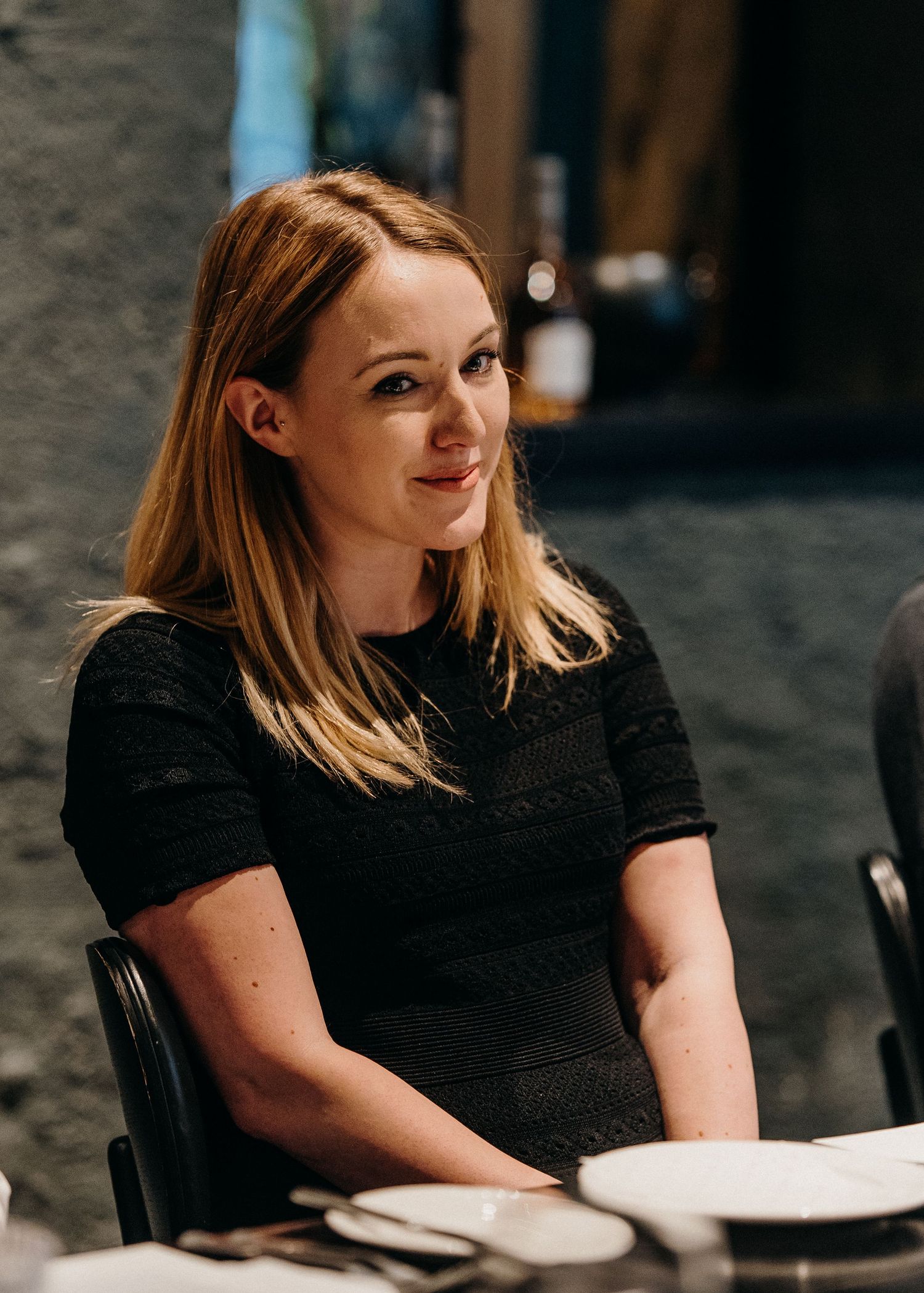 Woman with blonde hair wearing a black top sits at a table in a dimly lit setting.