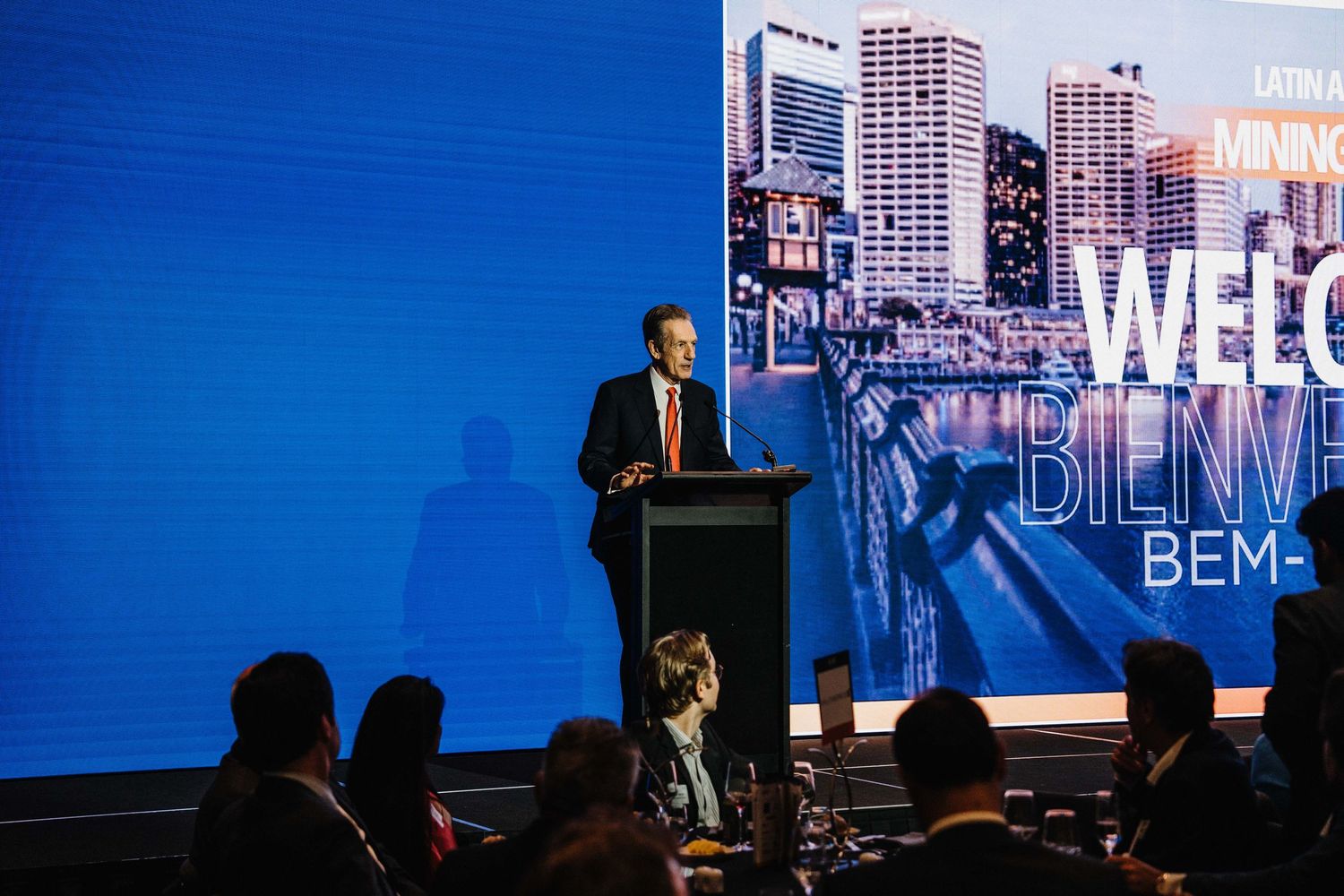 Business conference presenters speak on stage with blue lighting and large video screens displaying content.