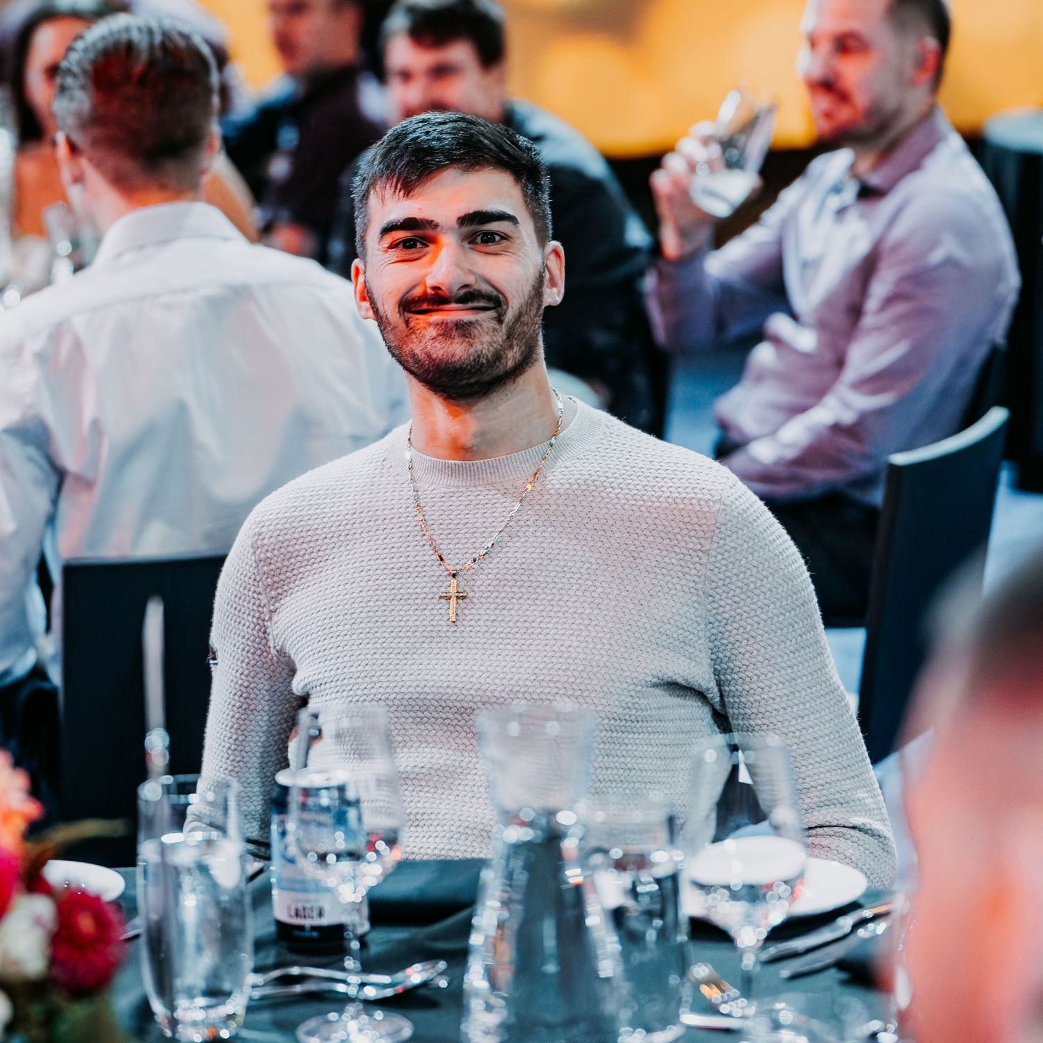 A person in a light colored sweater smiling at a formal dining event with glassware and table settings visible.
