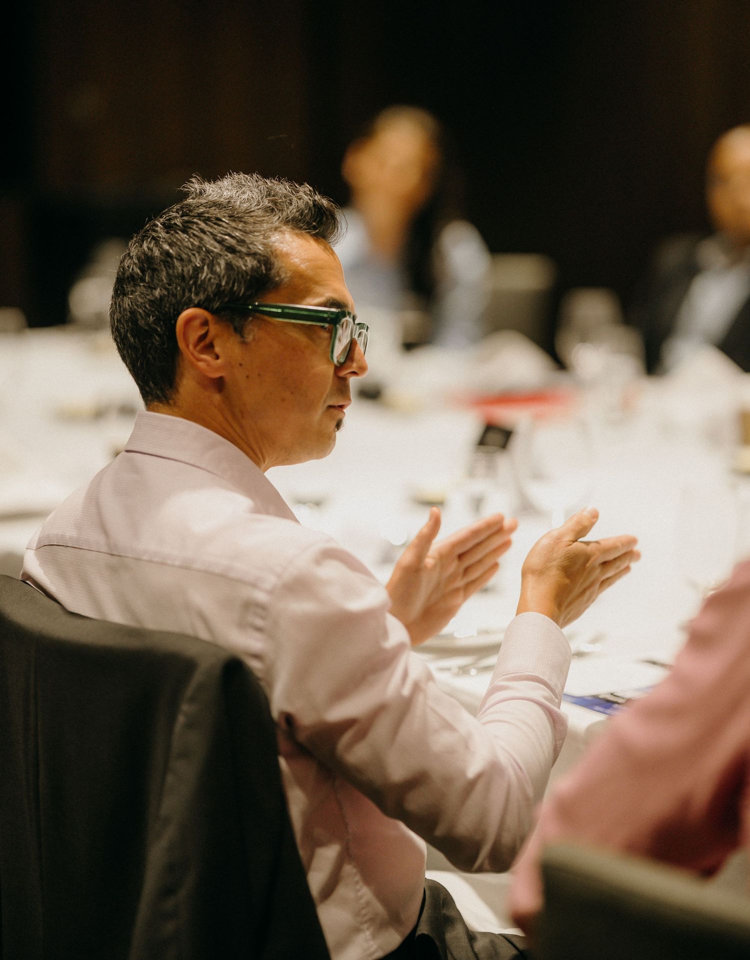 Man with glasses applauding at a formal dinner event, seated at a white tablecloth table in a dimly lit venue.