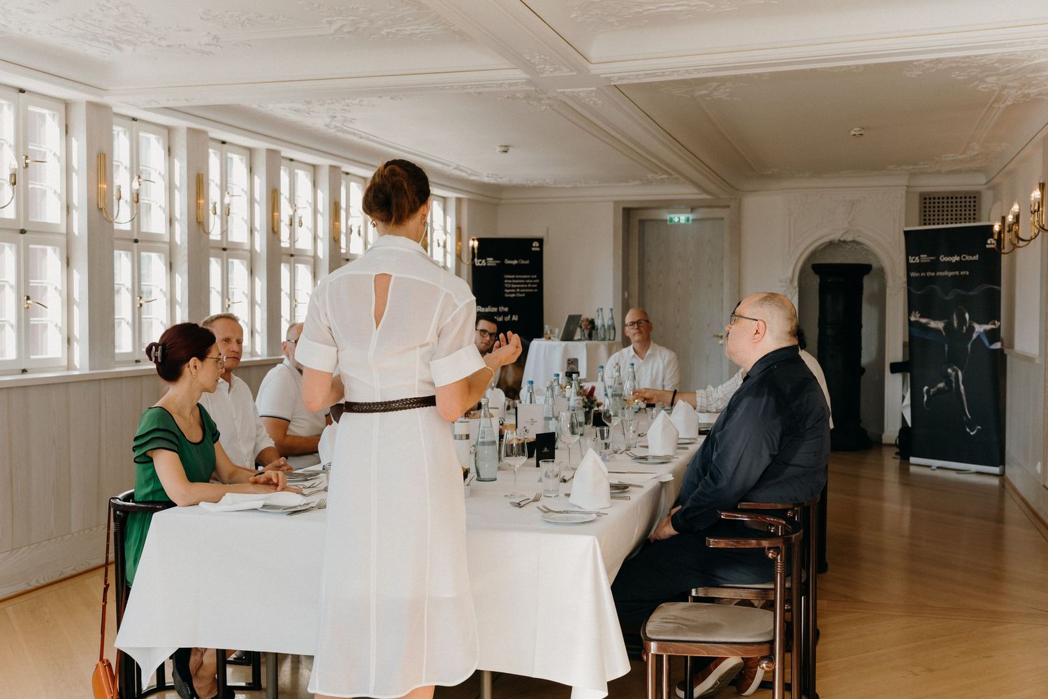 Woman in white dress addresses guests seated at elegant dining table in bright formal room.
