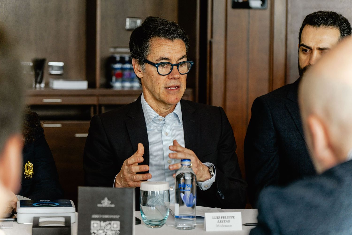 Business professional in glasses speaking at a corporate roundtable meeting with colleagues in a modern conference room.