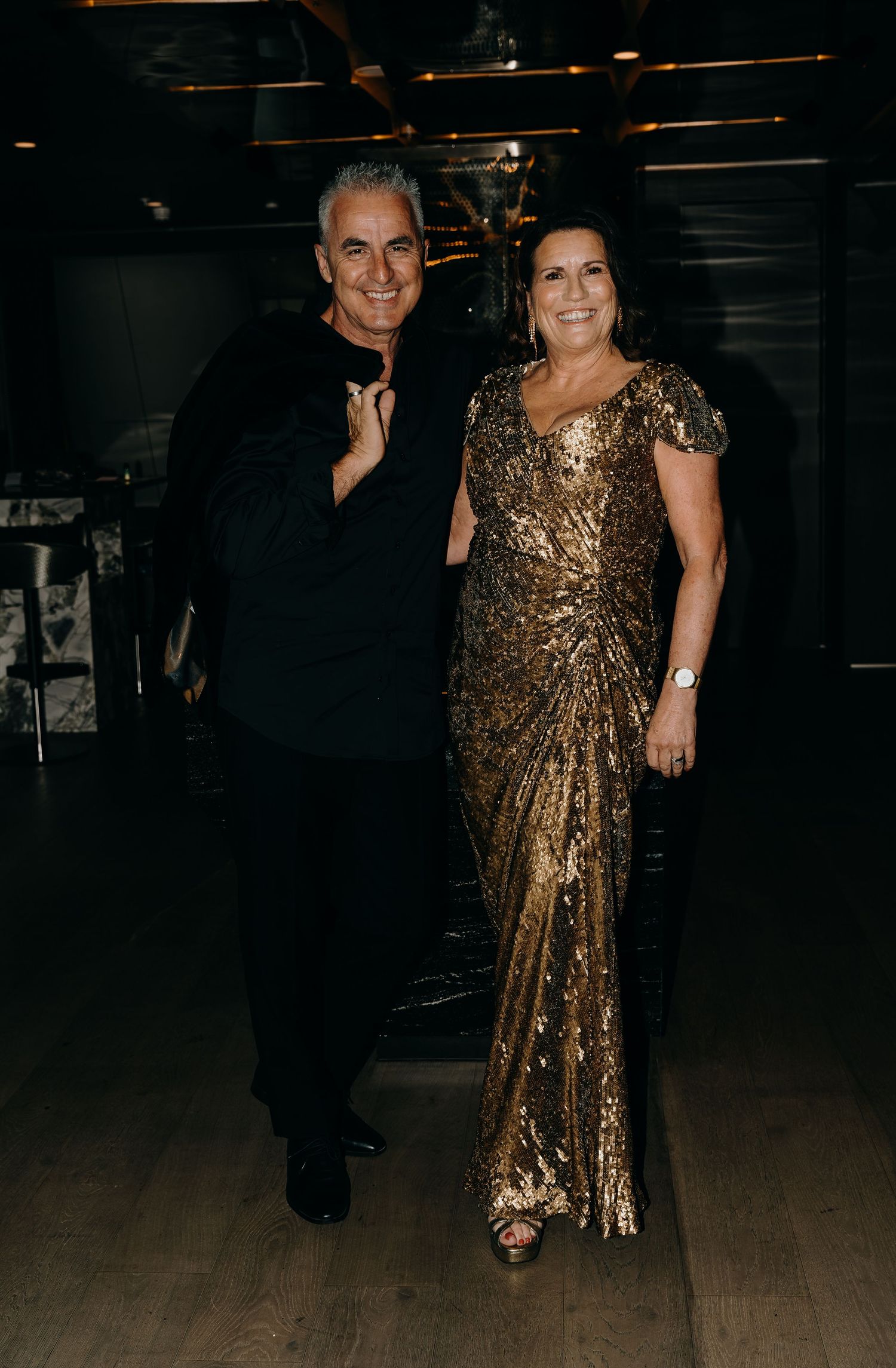 A couple in formal attire, with a sparkly gold dress and dark suit, dancing at an evening event indoors.