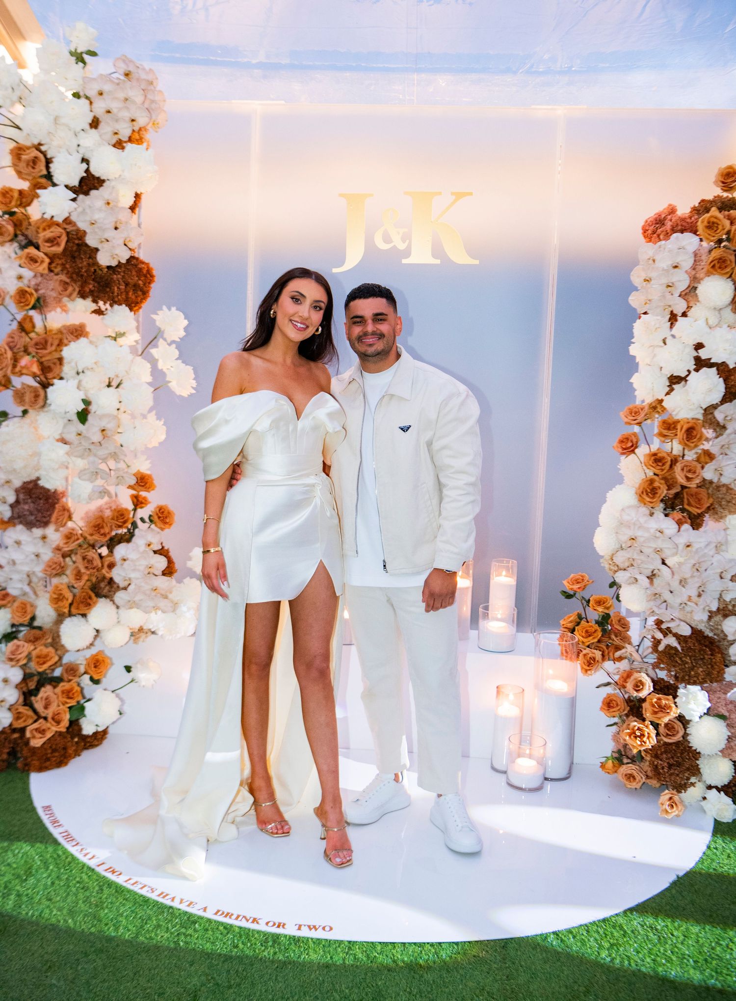 A couple sitting on a white circular platform surrounded by elegant floral arrangements of white and orange blooms.
