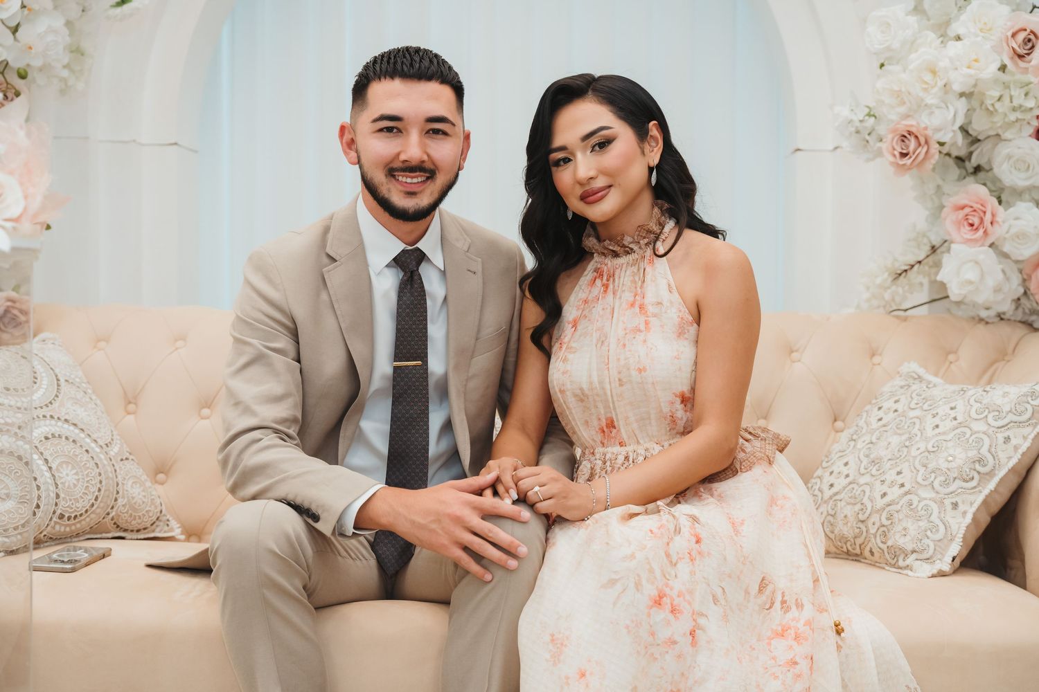 A romantic engagement photoshoot with soft floral decor and elegant formal attire against a light backdrop.