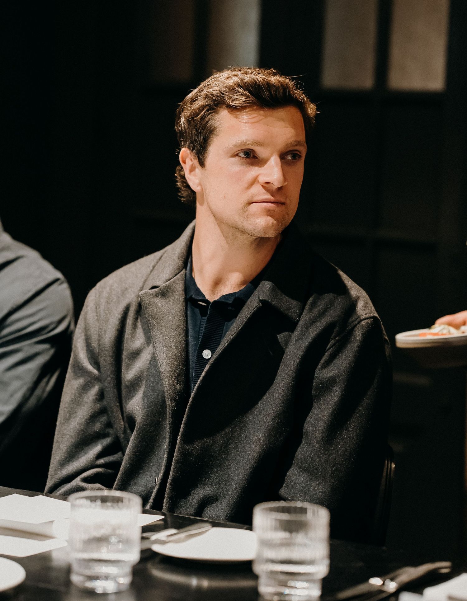 A person in a dark suit sits at a table with a glass of water in a dimly lit setting.