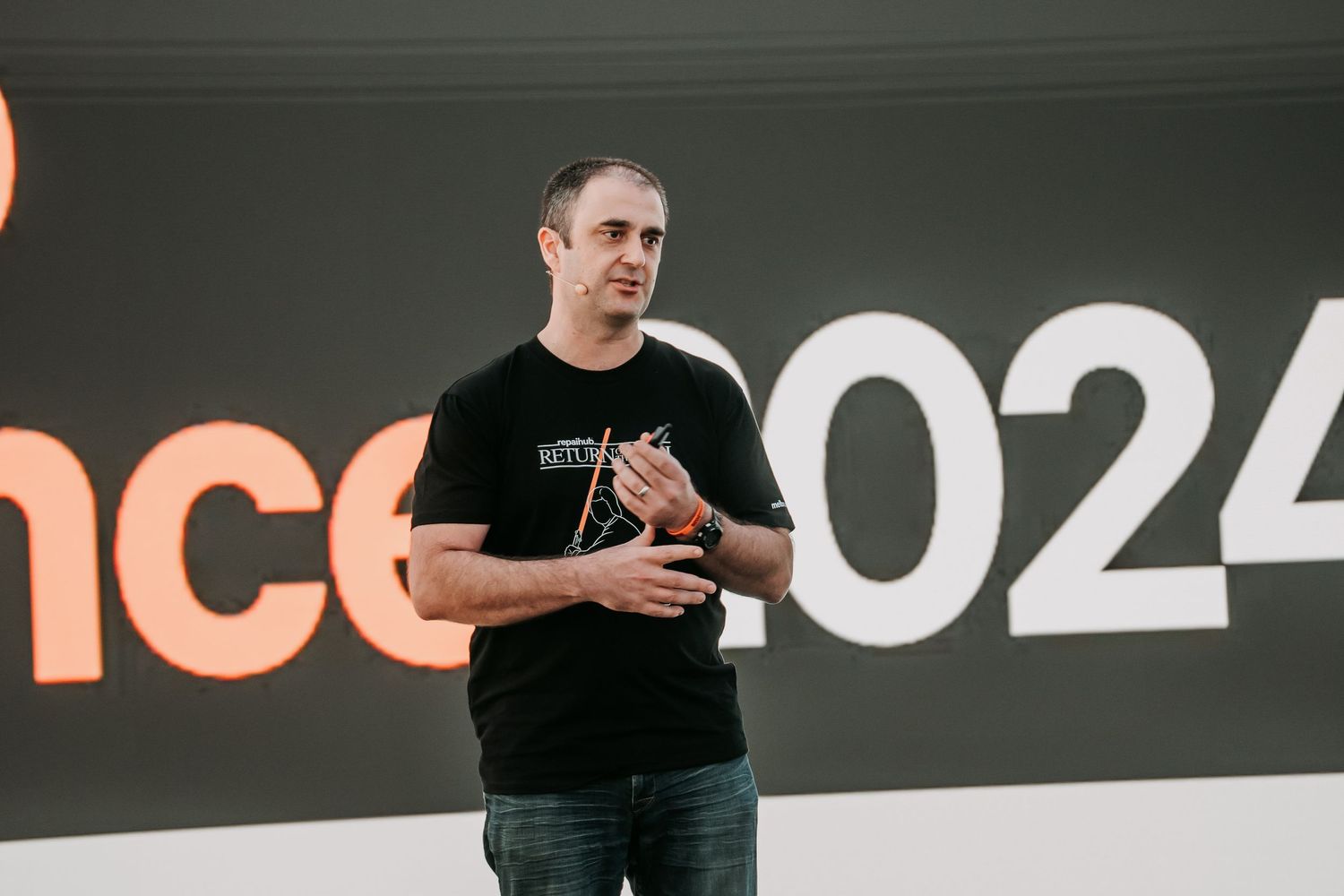 Person in a black t-shirt gestures while giving a presentation in front of a dark background with numbers.