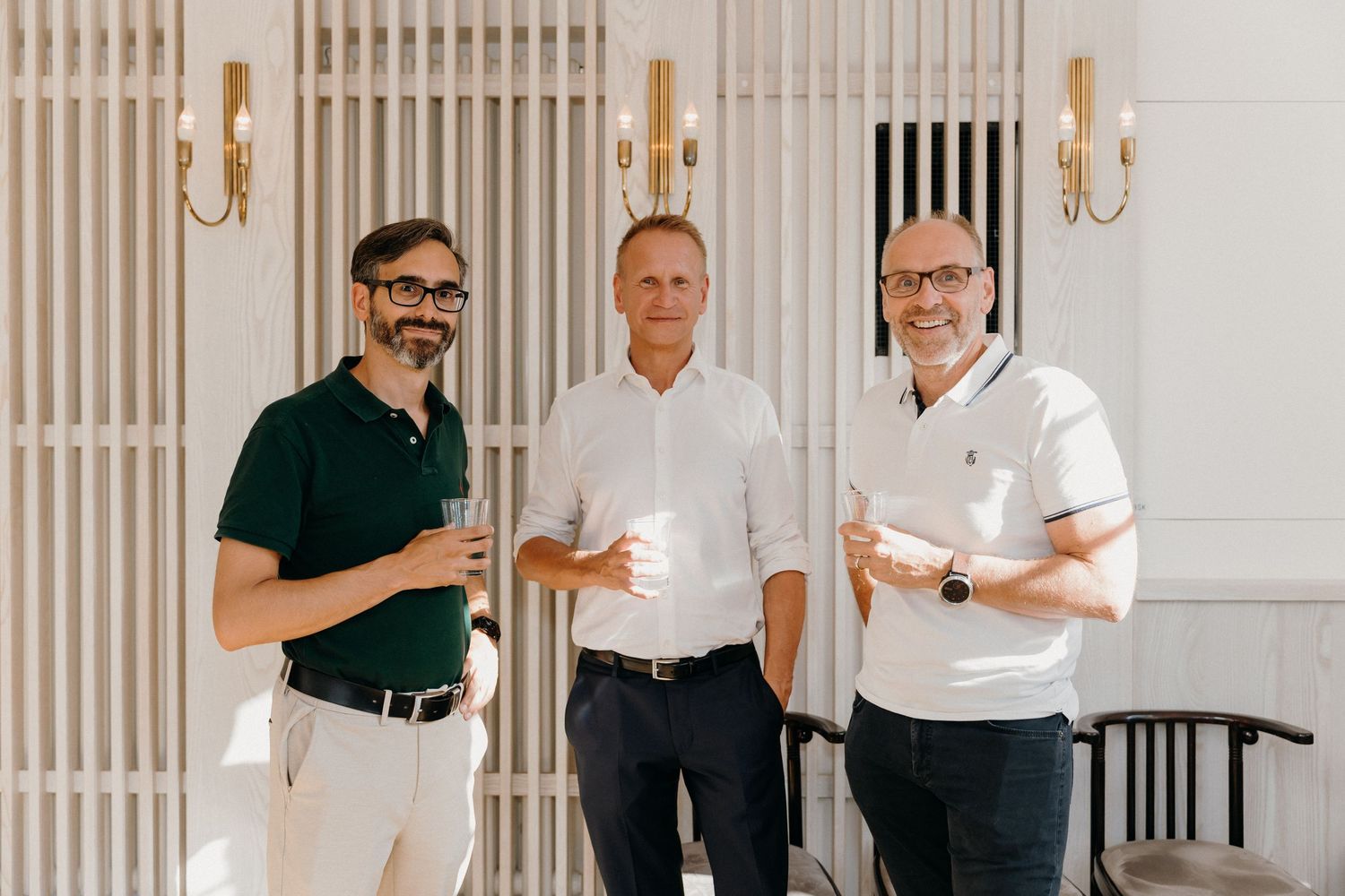 Three smiling men standing together in an elegant interior, holding coffee cups in a bright, modern setting.