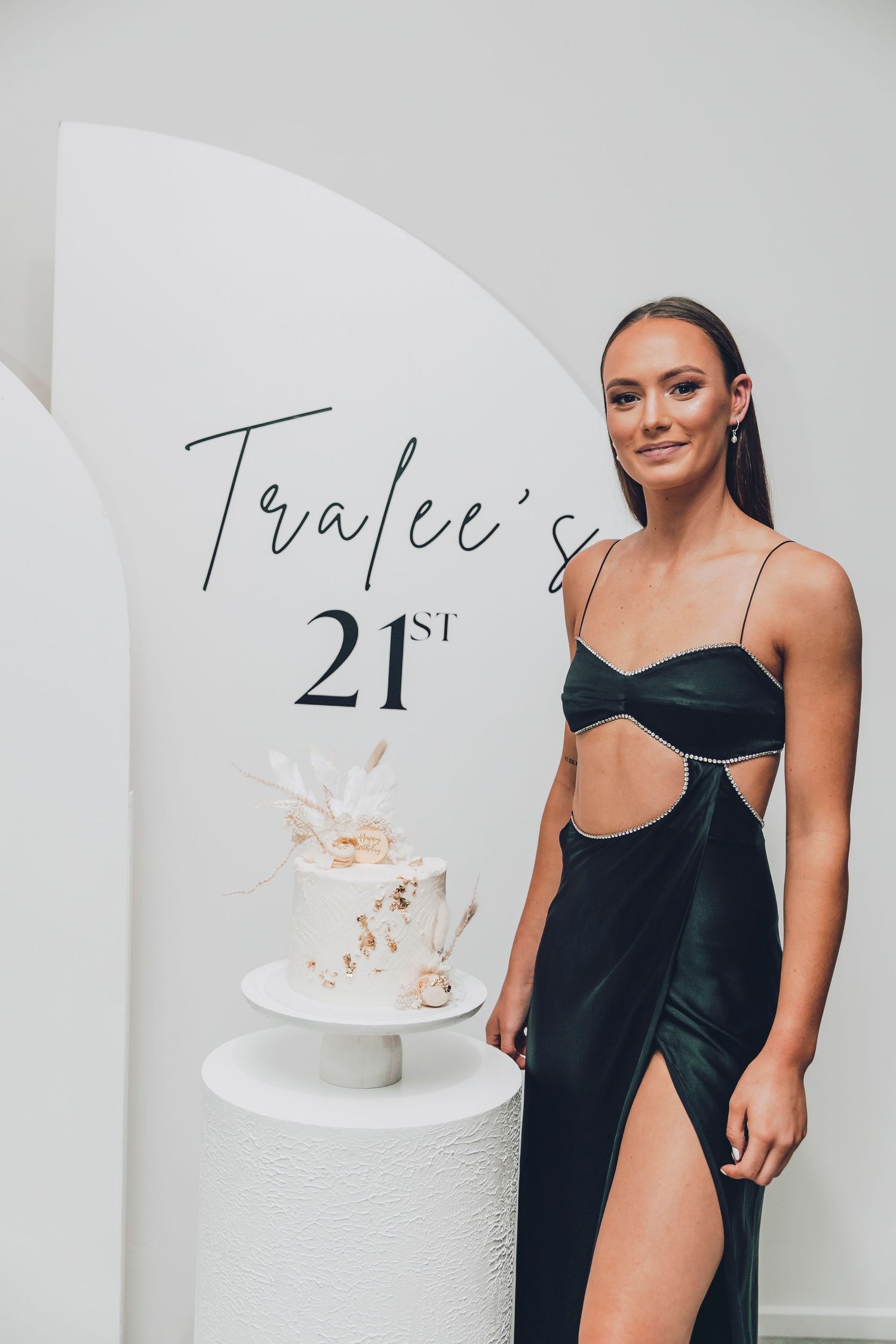 Woman in black dress posing with decorative white birthday cake against white background.