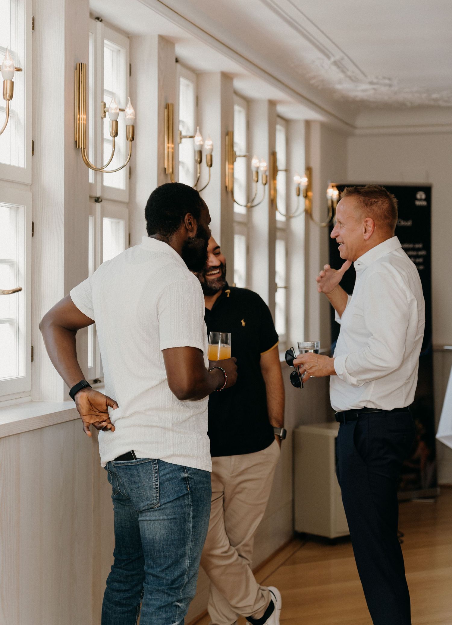 Three men laughing and conversing in an elegant room with gold wall sconces and large windows.
