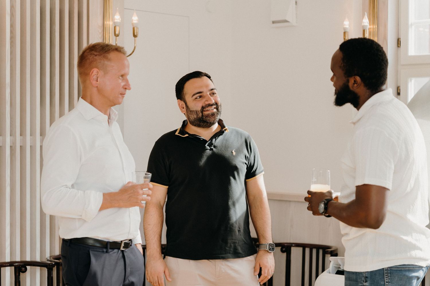 Three men smiling and chatting at a social gathering, holding drinks in a bright, elegant interior setting.