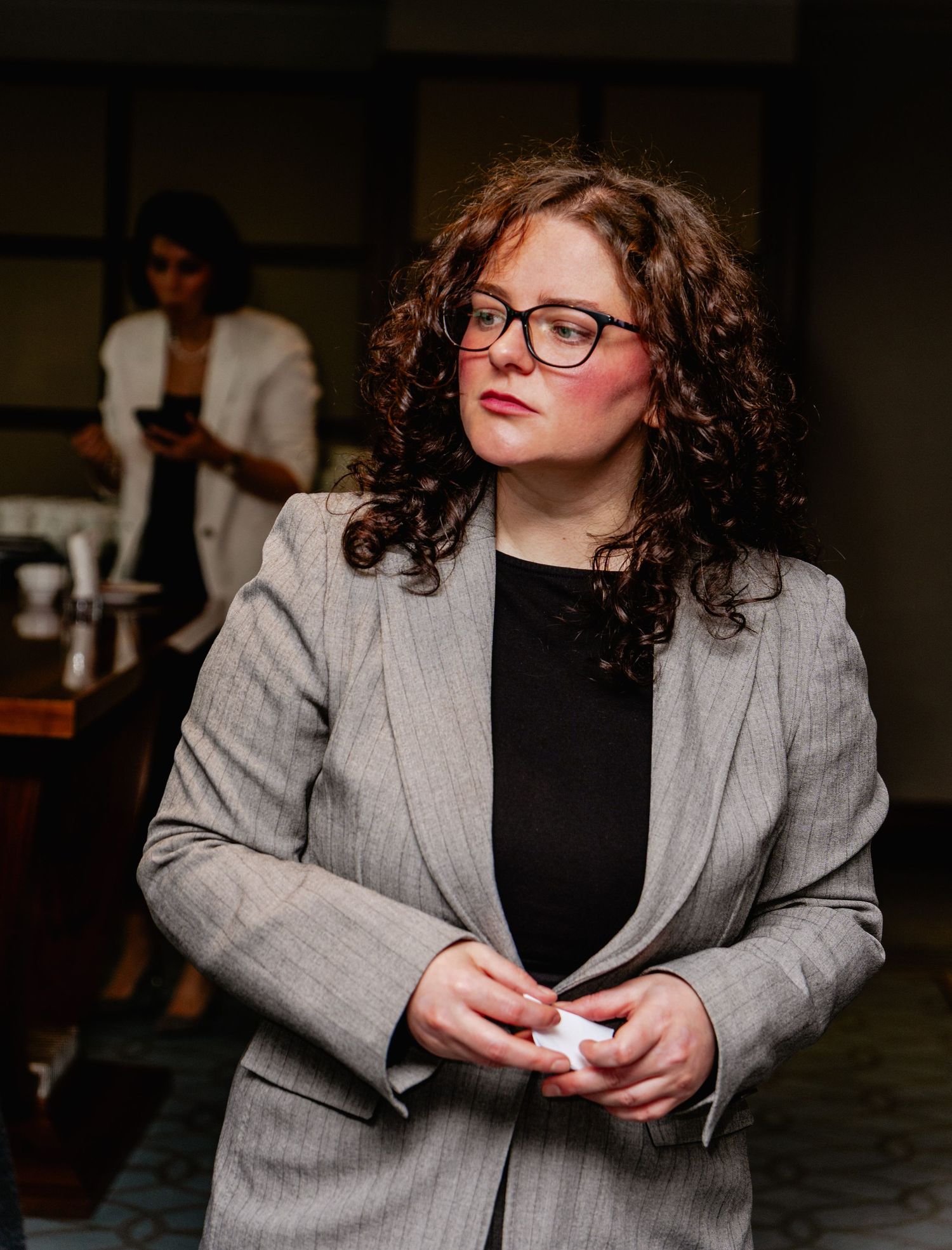 Woman with curly hair and glasses wearing a gray blazer stands indoors holding a small white object.