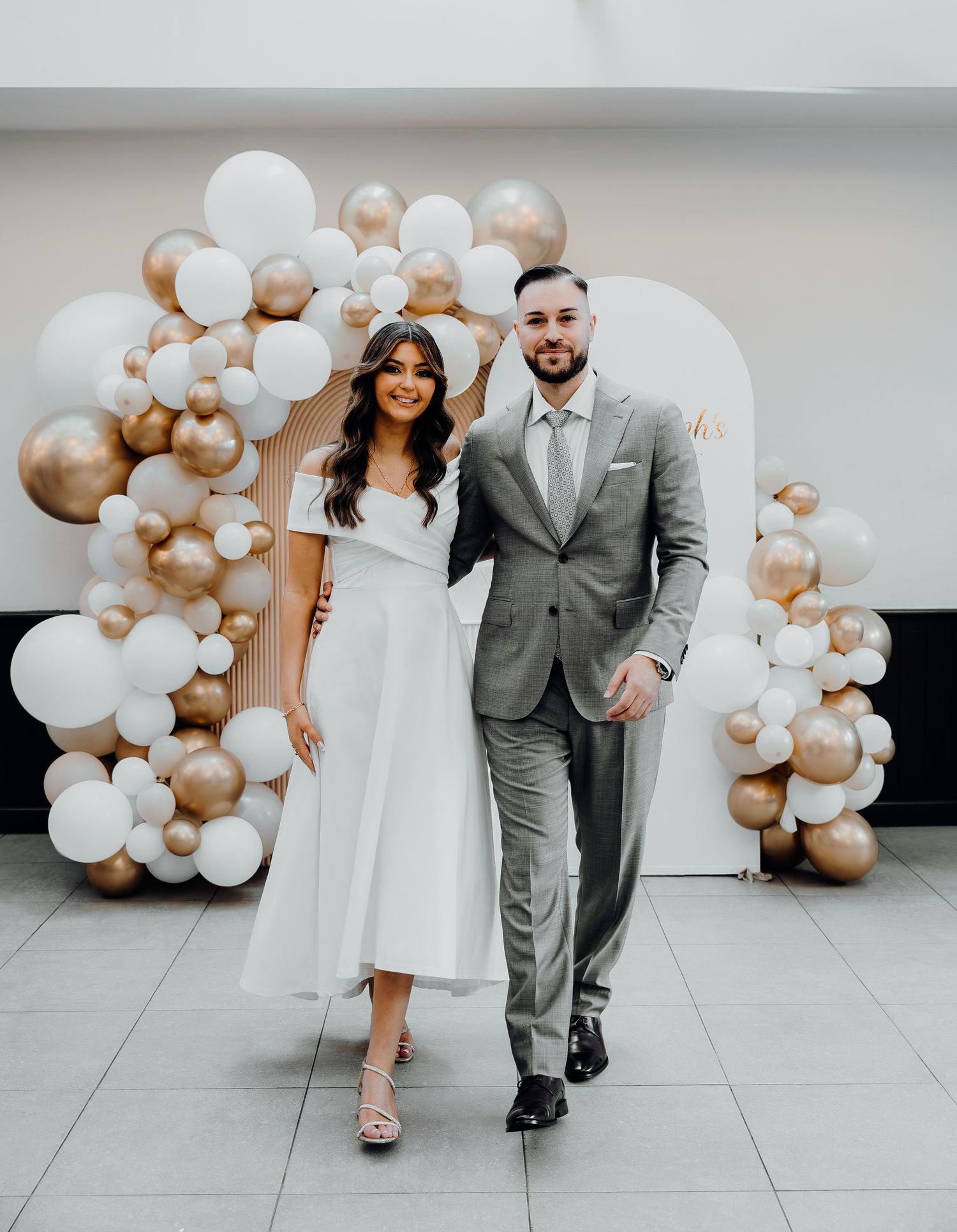A couple walks side by side under an elegant balloon arch with white and champagne colored balloons at an indoor venue.