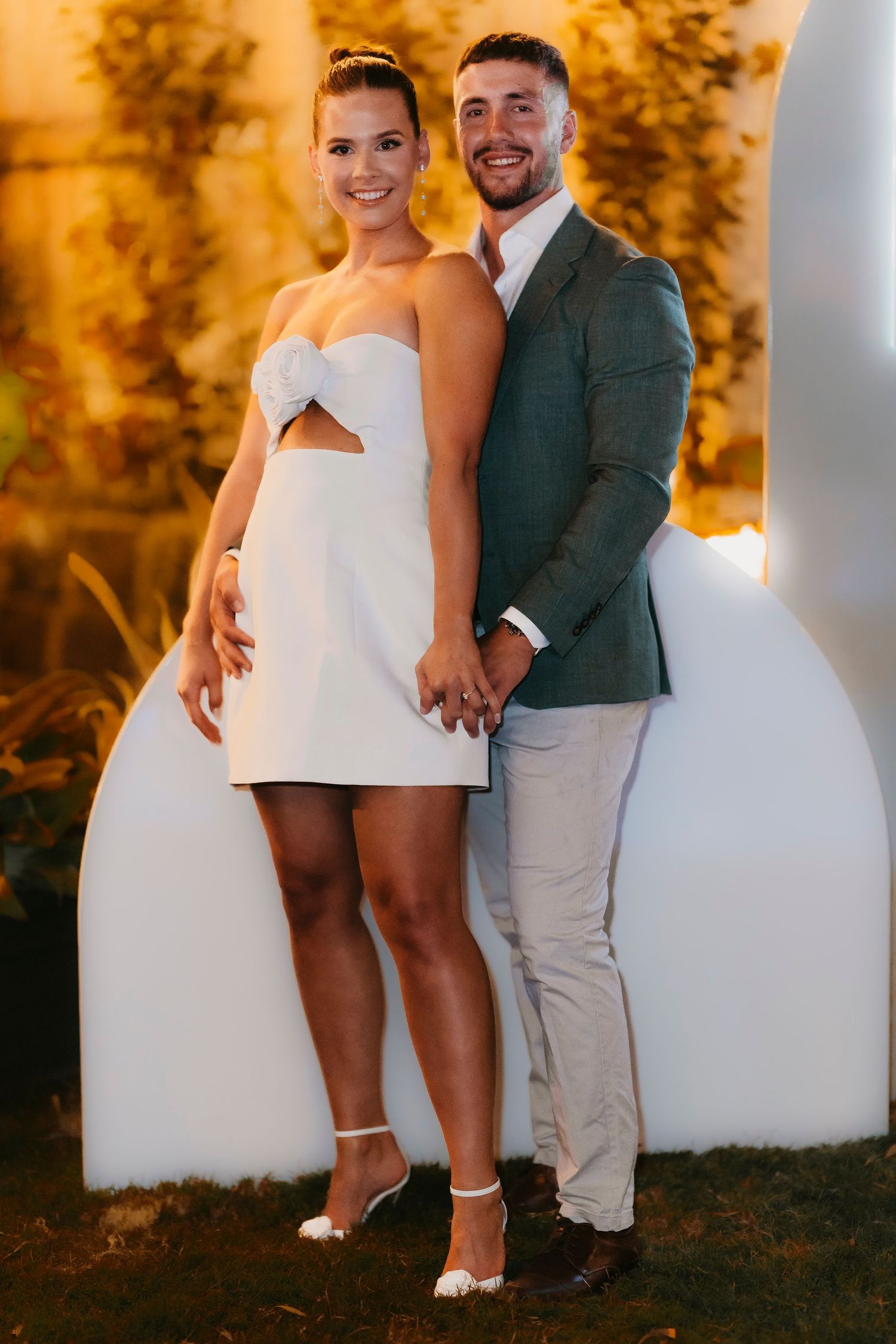 A couple poses together outdoors at sunset, creating intimate photos in warm autumn lighting against a white wall.