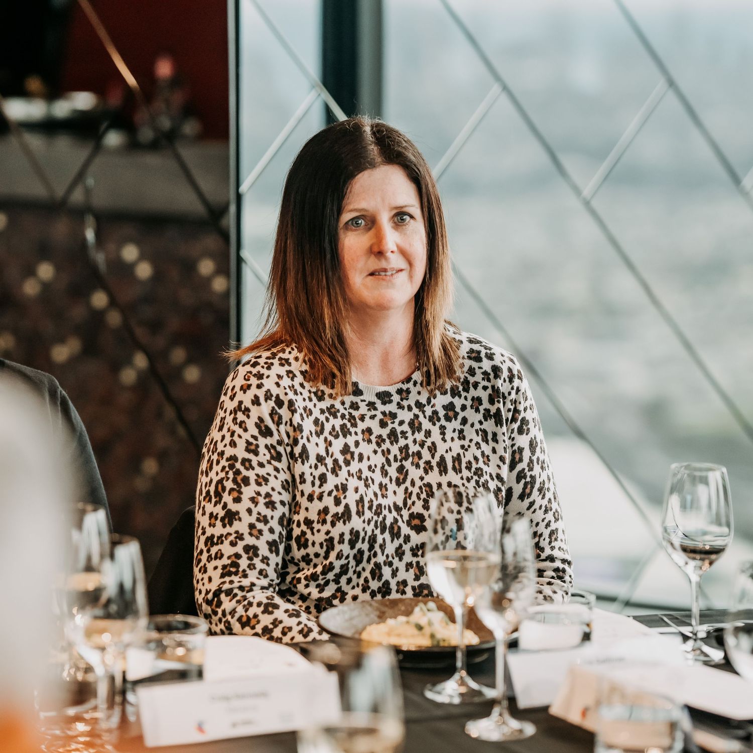 Person in leopard print top dining at upscale restaurant with wine glasses and place settings on table.