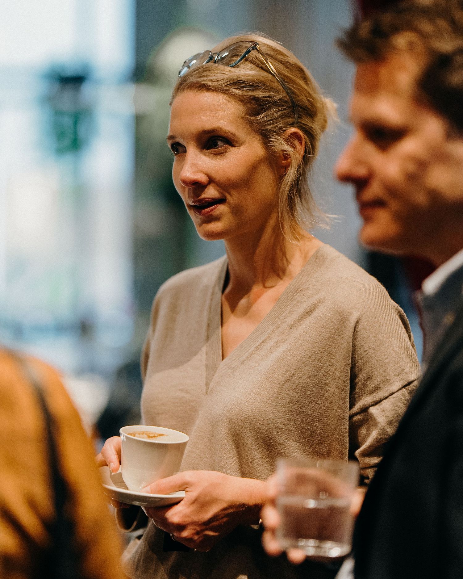 A person in a beige sweater holds a white coffee cup while conversing with others in an indoor setting.