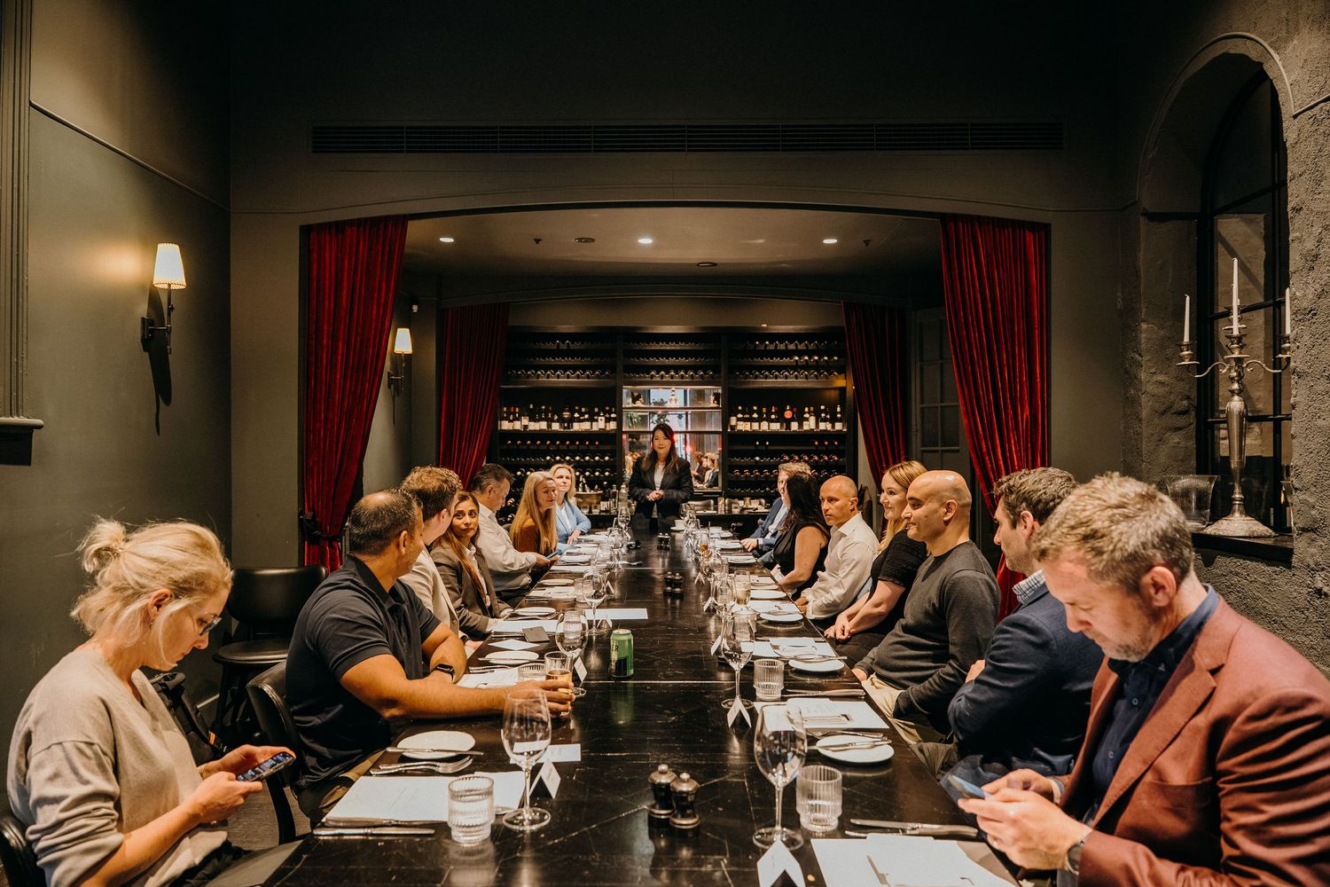 Guests seated at a long dining table in an elegant restaurant with red curtains and stone archways in warm lighting.