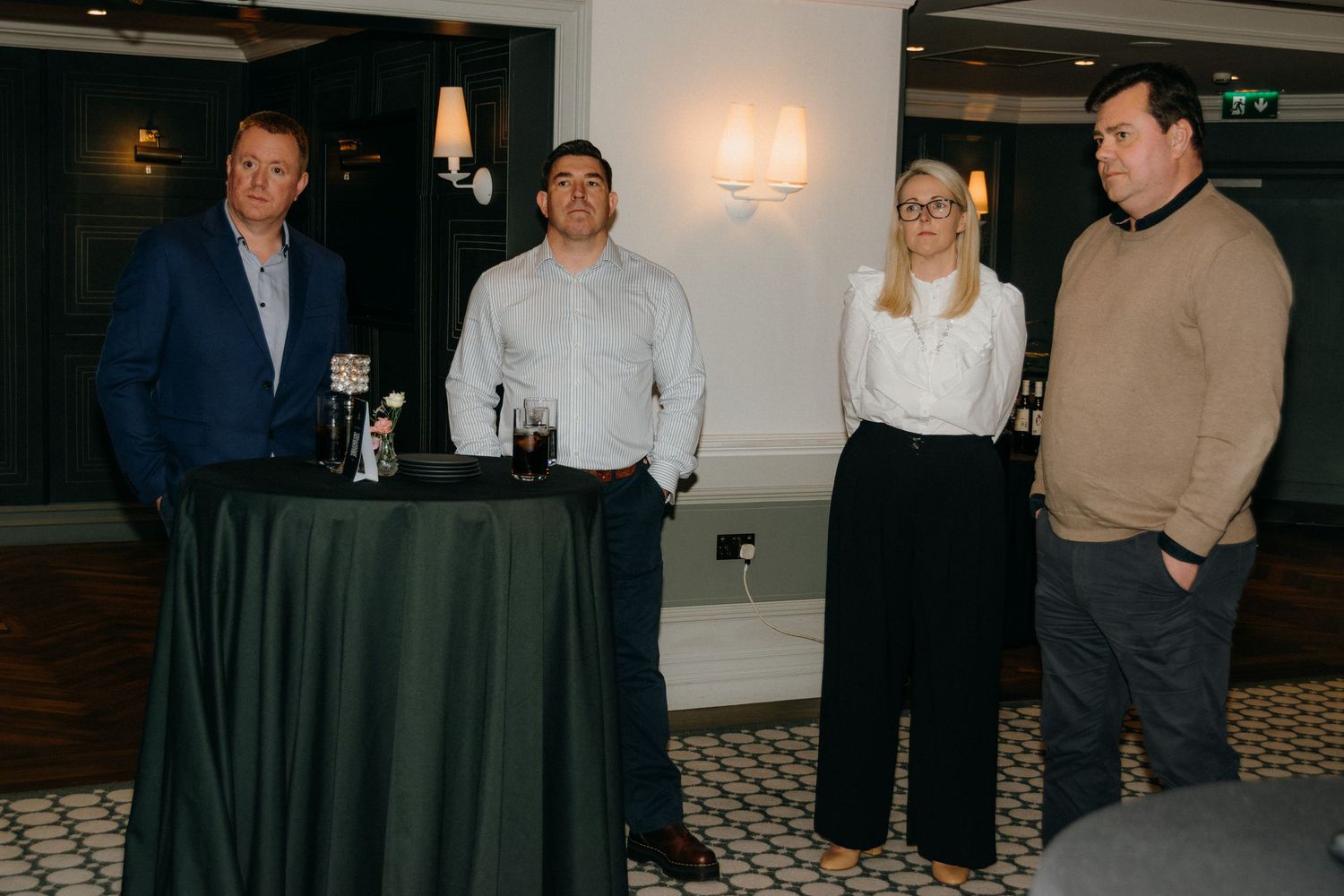Four people in business attire stand together at a cocktail event with a draped table in a dimly lit venue.