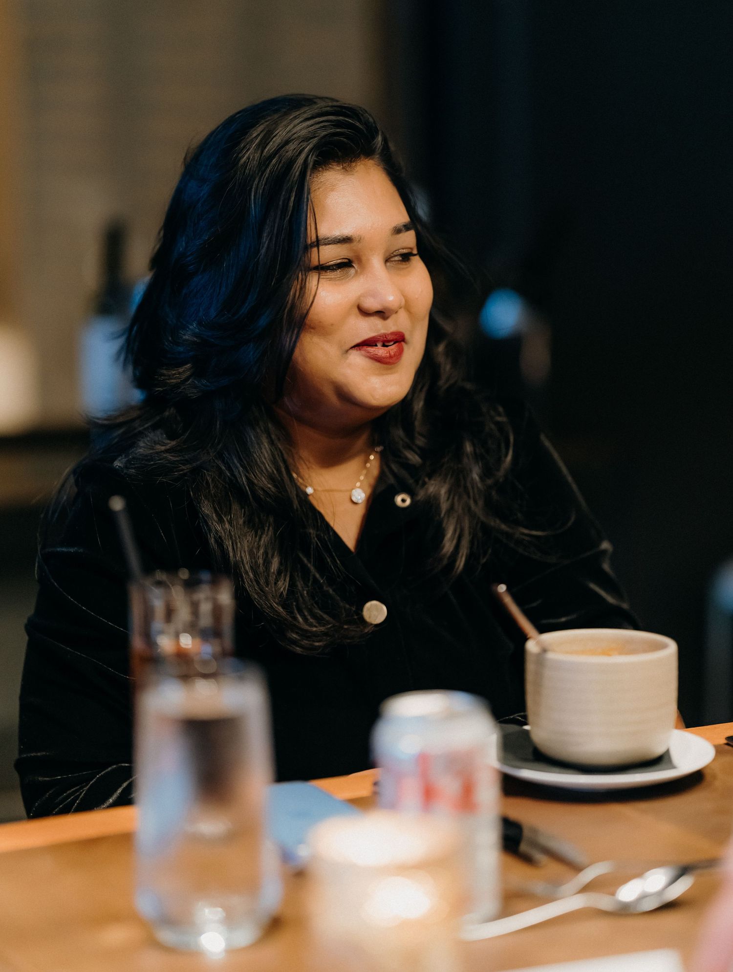Person in black attire sitting at a dining table with coffee cups and glassware in a dimly lit restaurant setting.