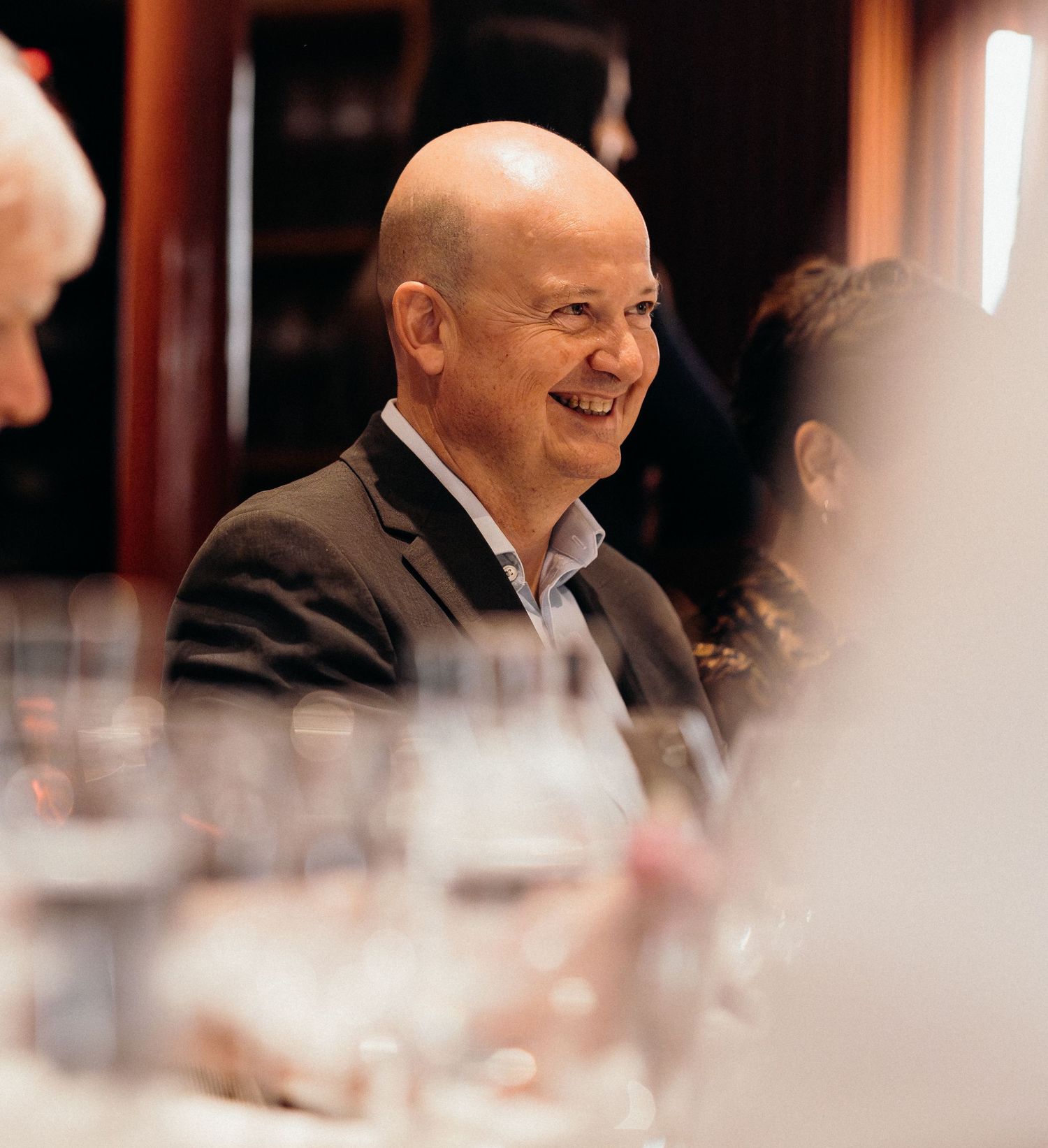 A smiling person in a dark suit jacket sits at a dining table with wine glasses in the foreground.