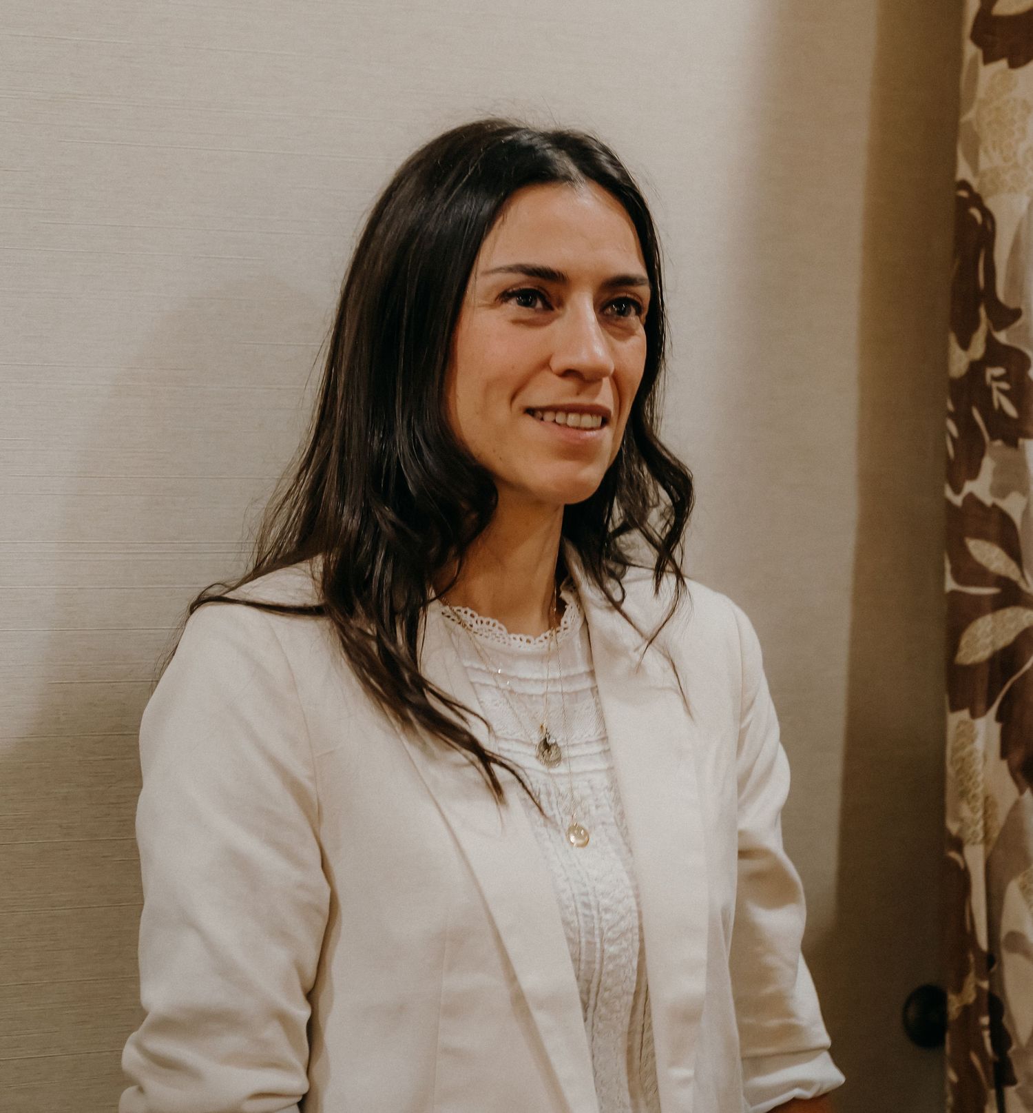Person with long dark hair wearing a white sweater smiles while looking to the side in an indoor setting.