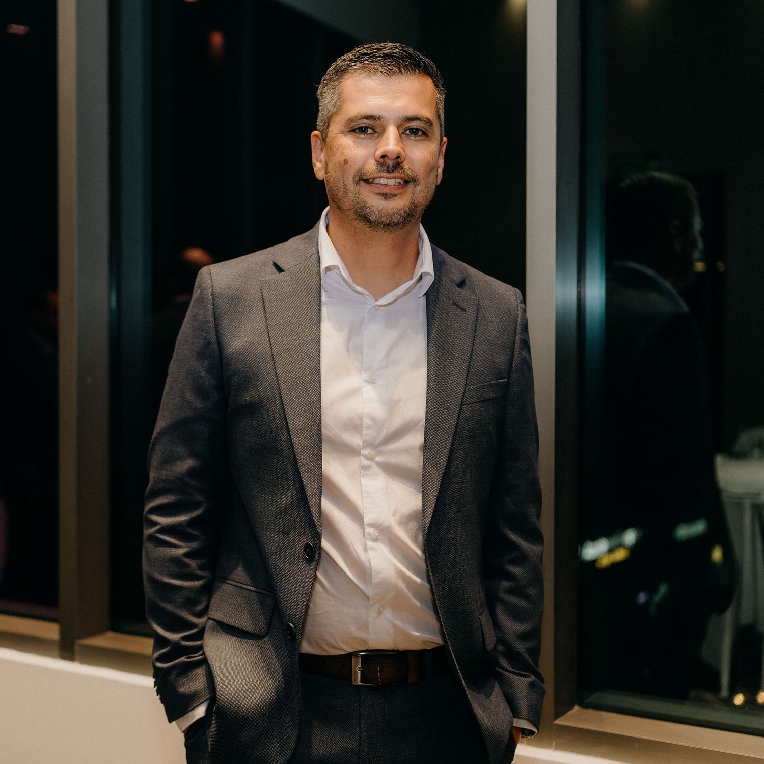 Professional in a dark suit and white shirt standing in a modern office setting with glass walls and dim lighting.