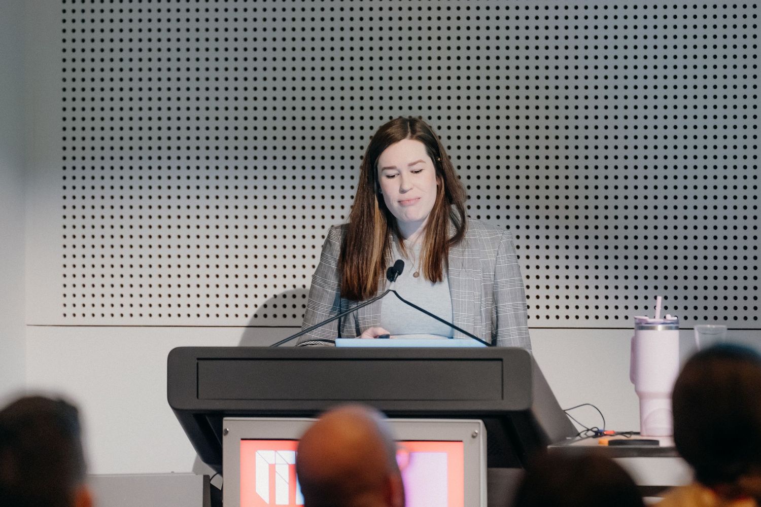 A person speaks at a podium during a conference presentation with perforated wall panels visible in the background.