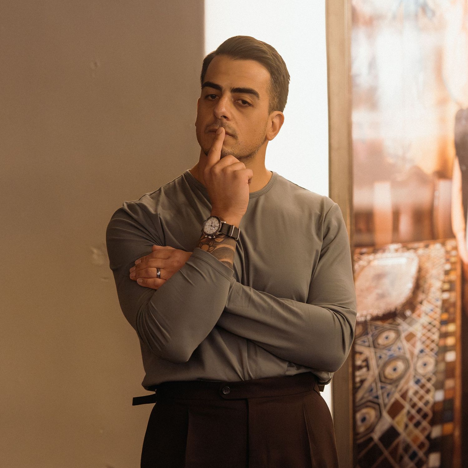Person in gray sweater standing thoughtfully against wall with patterned decor in warm lighting.