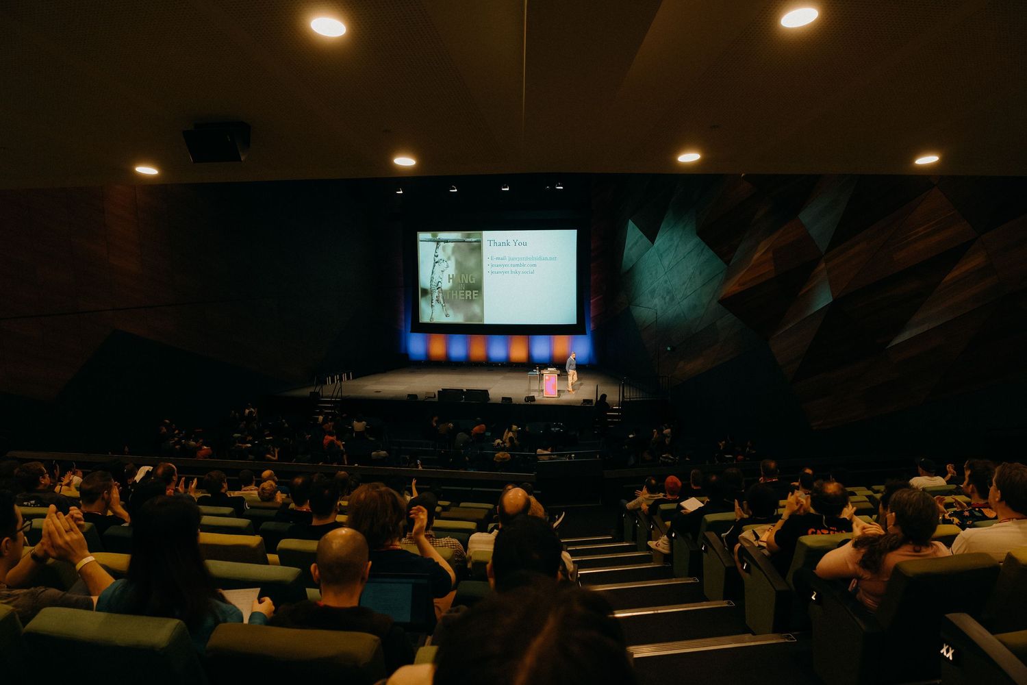 Dimly lit auditorium with rows of people seated during a presentation or lecture.