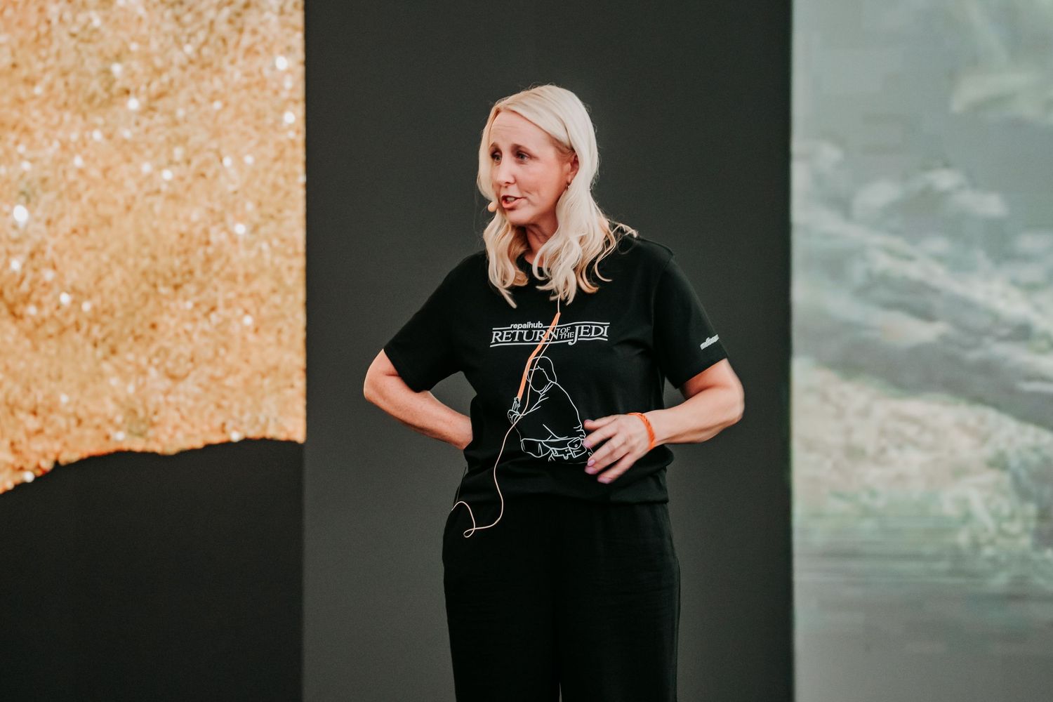Speaker wearing a black outfit presents on stage with hands on hips against a textured background.