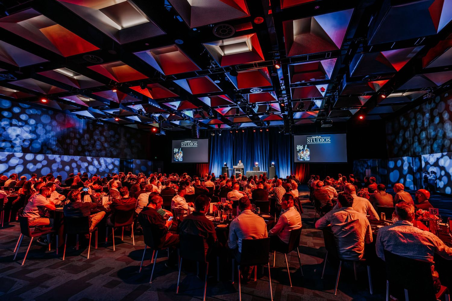 Panoramic view of a dimly lit conference hall filled with attendees seated at round tables during an evening event.