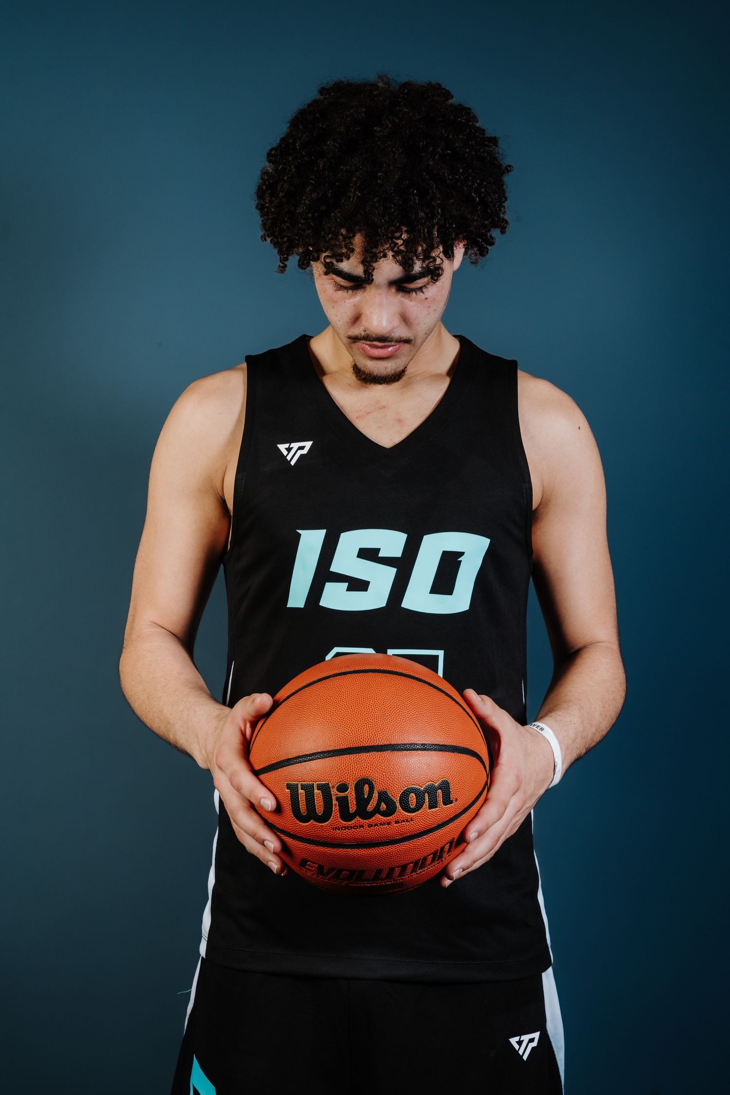Basketball player in black ISO jersey holds Wilson basketball against dark blue studio background.