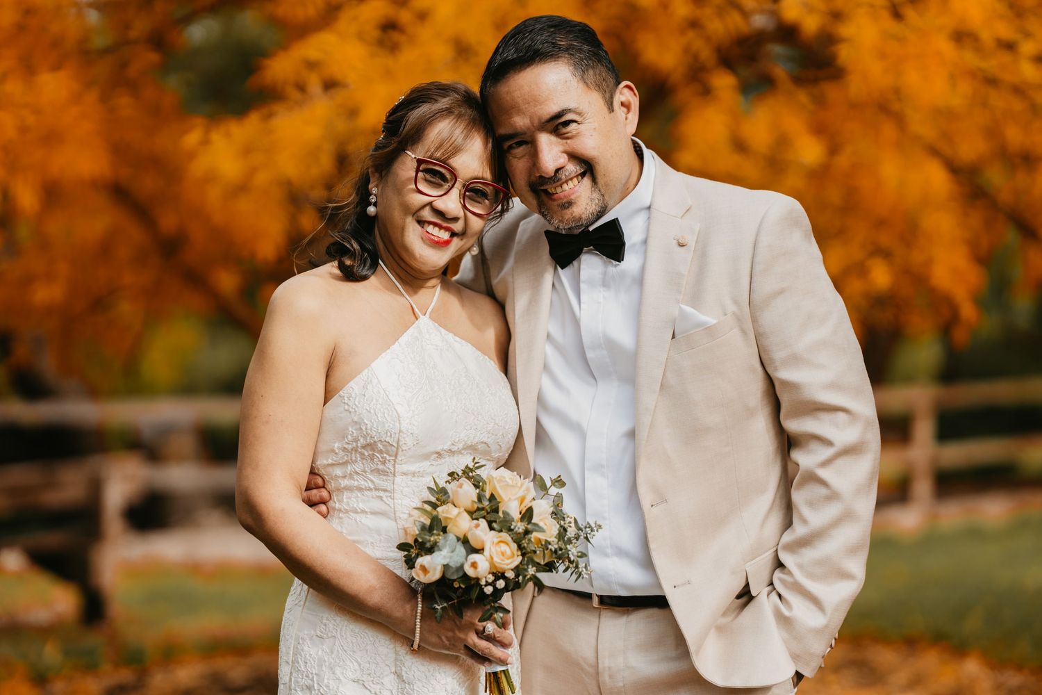 A couple shares a tender moment outdoors against vibrant autumn foliage in wedding attire.