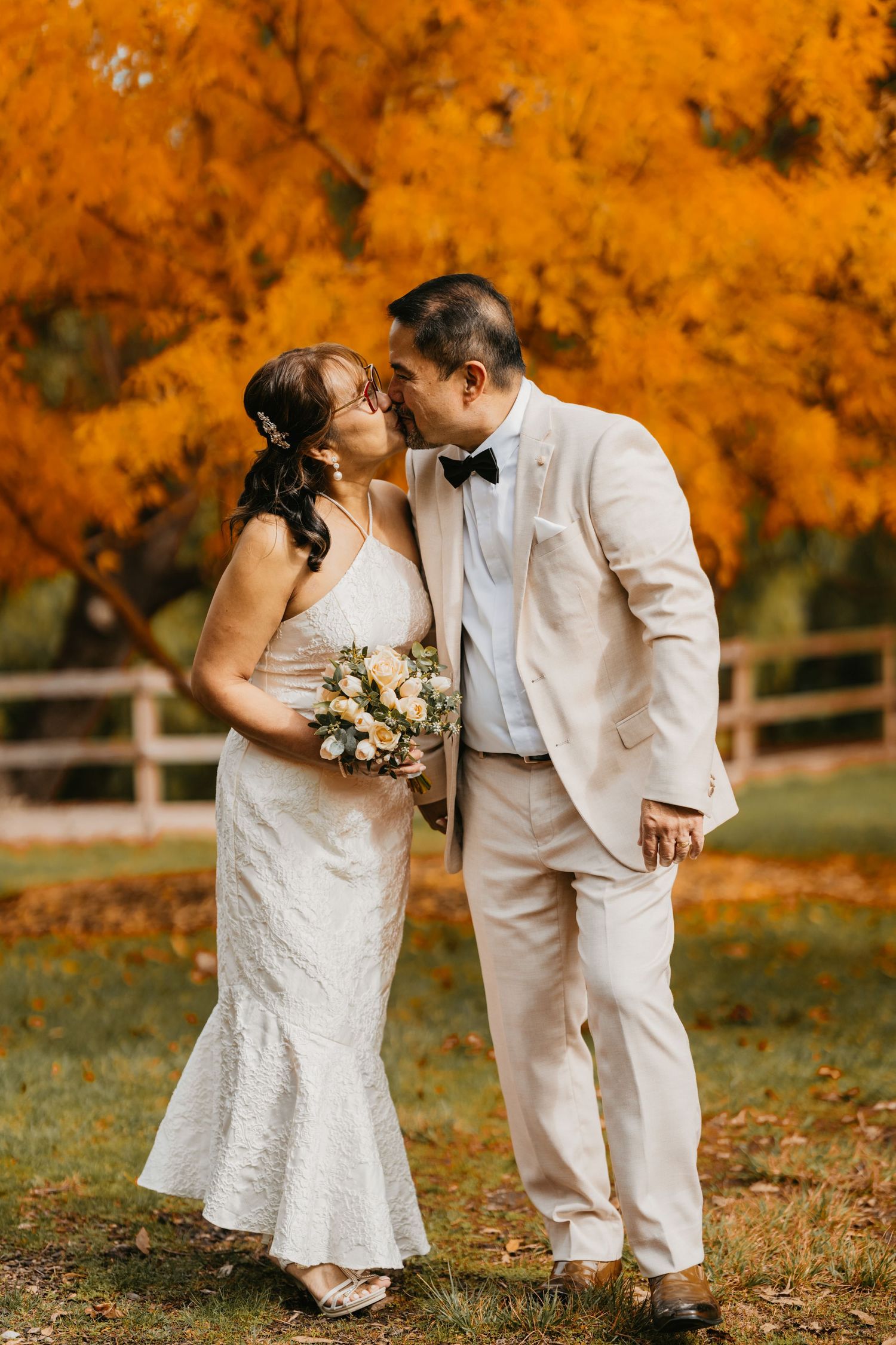 A couple in white wedding attire shares intimate moments against vibrant autumn foliage in a romantic outdoor setting.