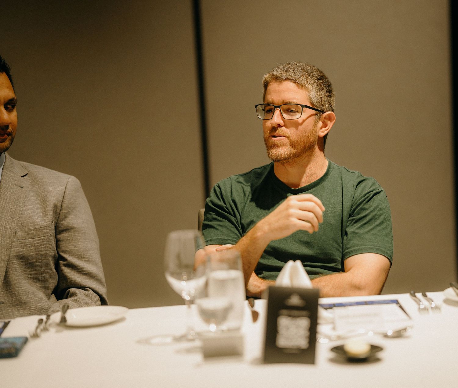 Two men seated at a formal dining table during a business dinner event, one wearing a green t-shirt and glasses.