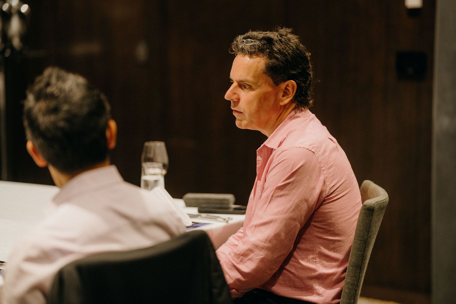 Two men in business casual attire seated at a dining table during a formal meeting or dinner event.