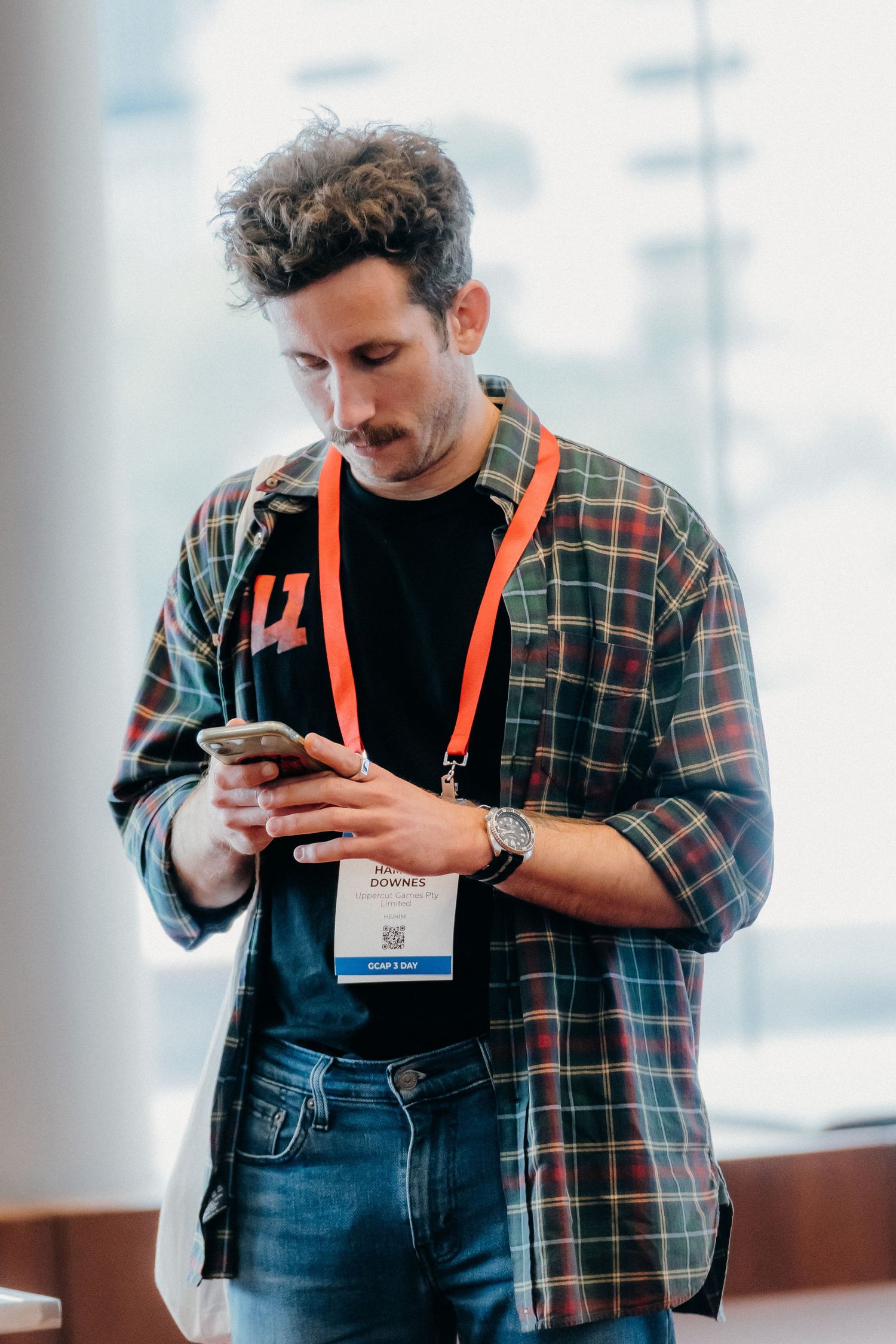 A person in a plaid shirt and overalls looks down at their mobile phone while wearing a conference badge lanyard.