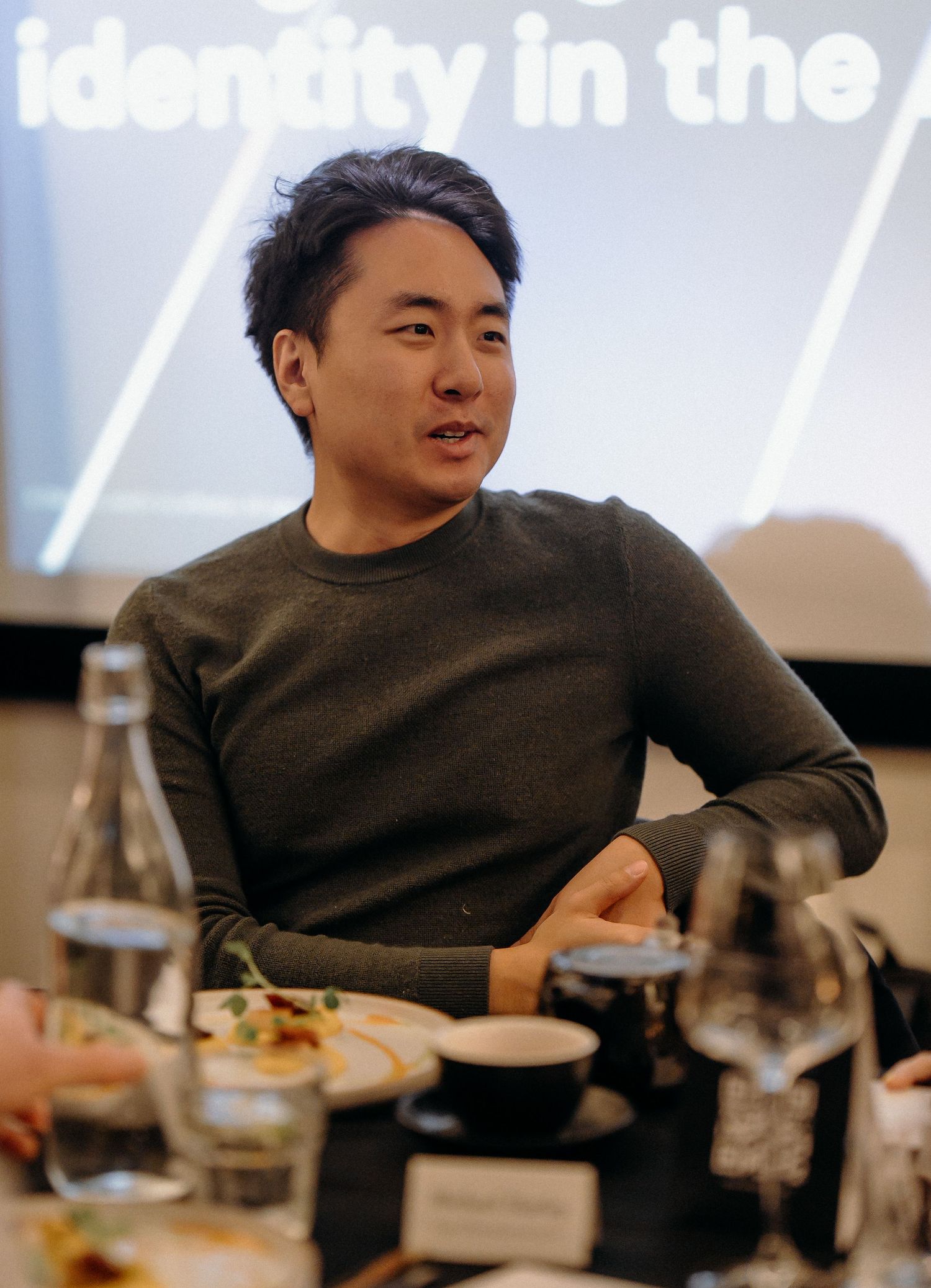 A person in a dark sweater sits at a dining table with plates and glasses during a meal.