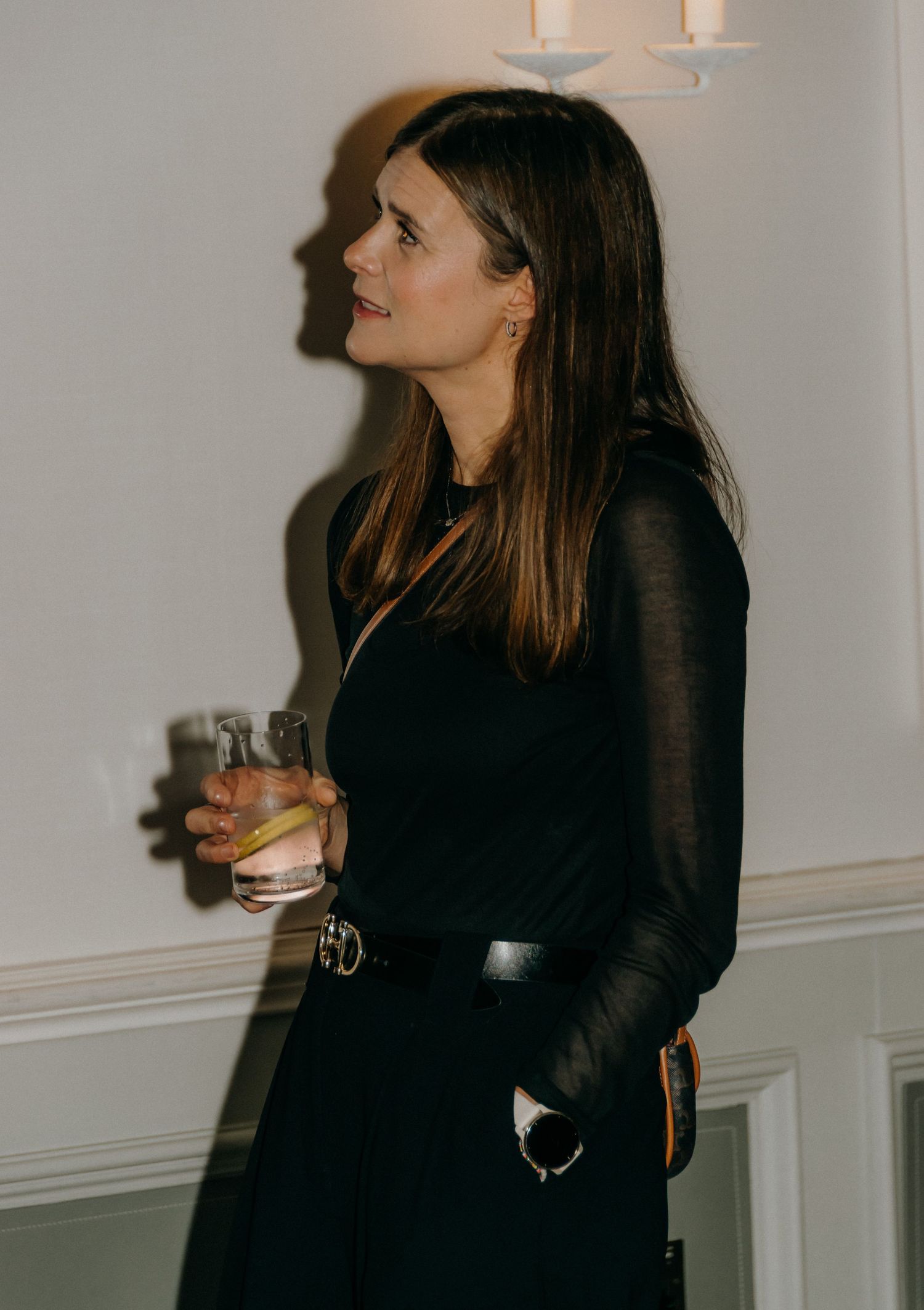 Woman in black long sleeve top leaning against wall while holding a beverage in dim lighting.