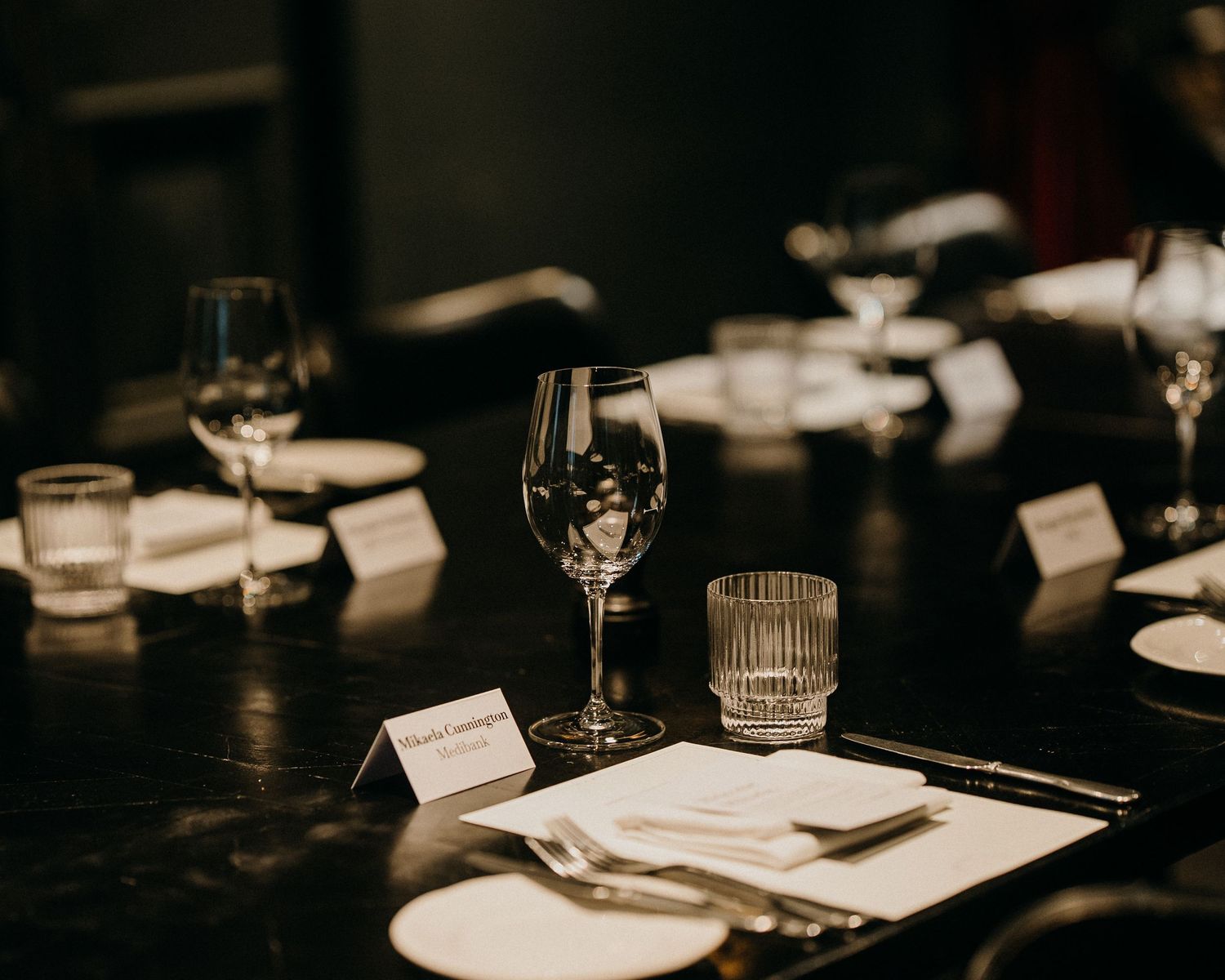 Place settings at an elegant dinner party with wine glasses and name cards on a dark table in moody lighting.