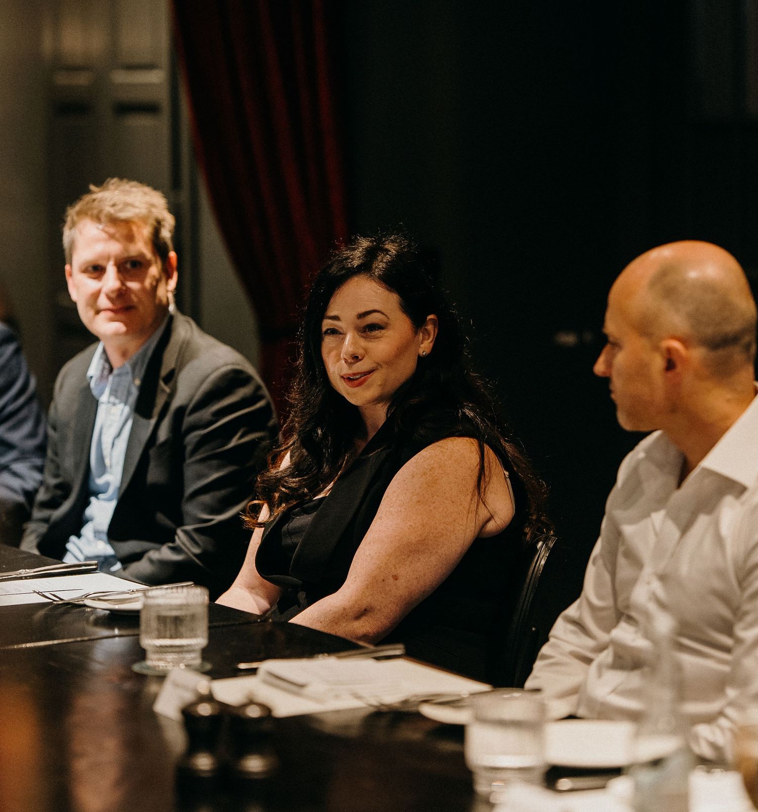 Three professionals seated at a conference table during a business meeting in a dimly lit room.