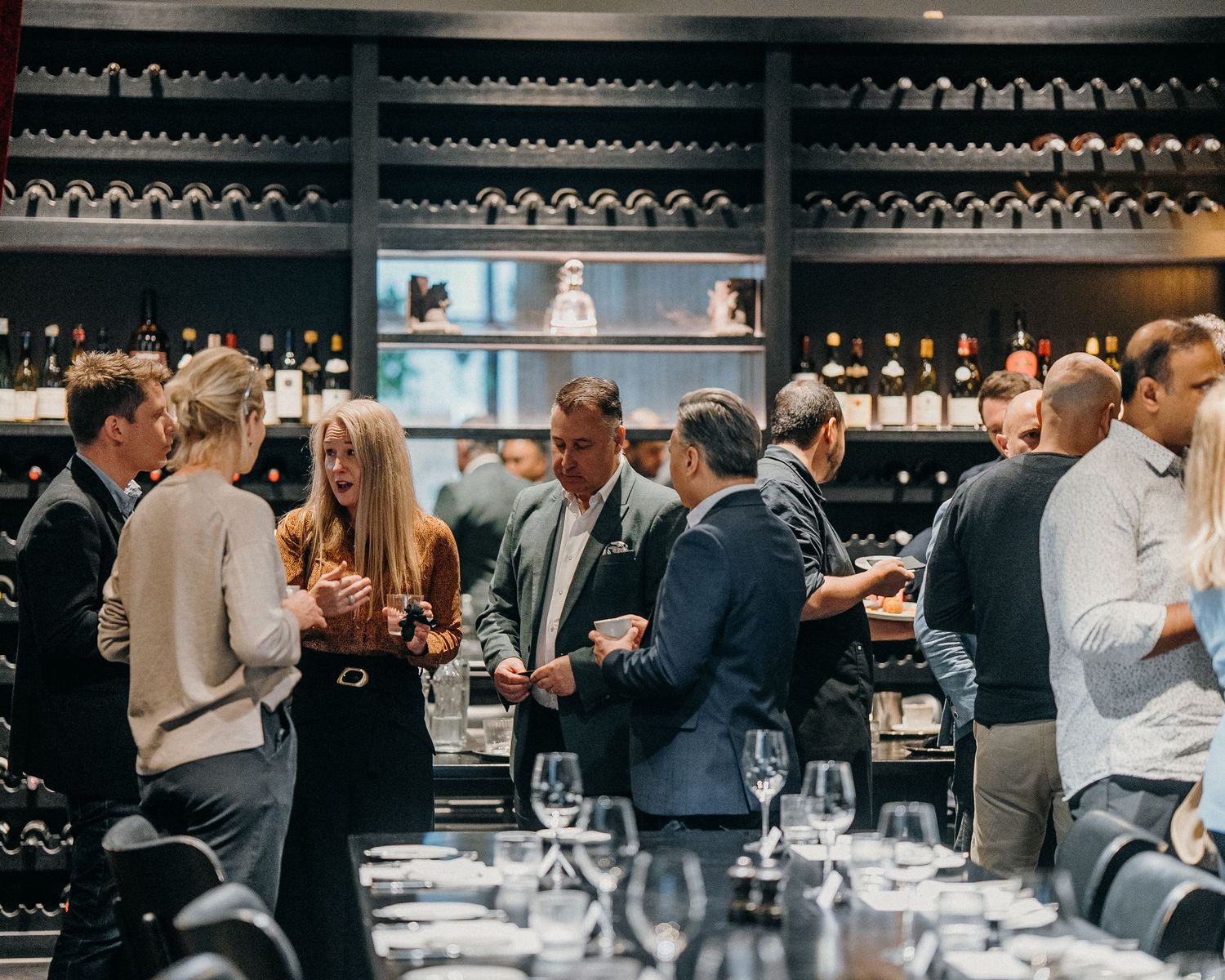 People gathered at a wine tasting event in a dark-lit room with bottles displayed on shelves in the background.